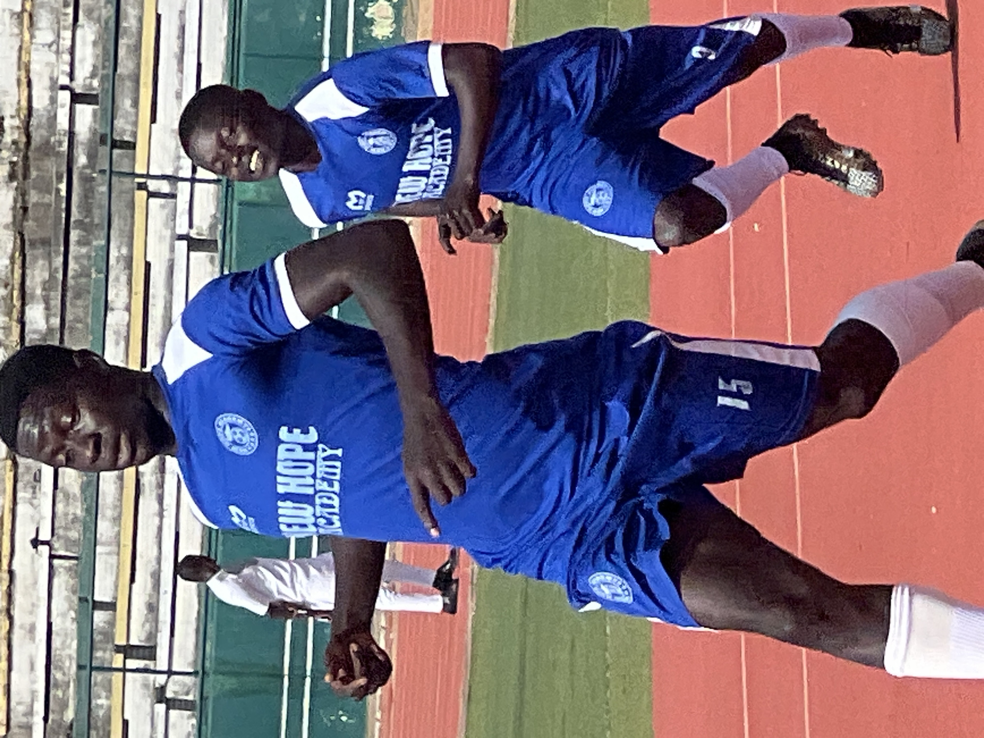 The image shows two young men playing soccer on a track and field at the New Stadium in Nigeria. They are wearing blue soccer uniforms with white trim and white socks. The man in the foreground is wearing shorts with the number 15 on them, and the man behind him has the number 13 on his jersey. Both men are dark-skinned and appear to be in good physical condition. The game seems to be intense, as the man in the foreground has his mouth open and looks focused on the game. The man behind him is also looking down at the ball with a determined expression. The stadium appears to be relatively empty, with only a few spectators visible in the background. The weather seems to be warm and sunny, as the players are sweating and the sun is shining brightly. There is visible text on the jerseys of the players. The text on the front of the jersey reads "NEW HOPE ACADEMY".