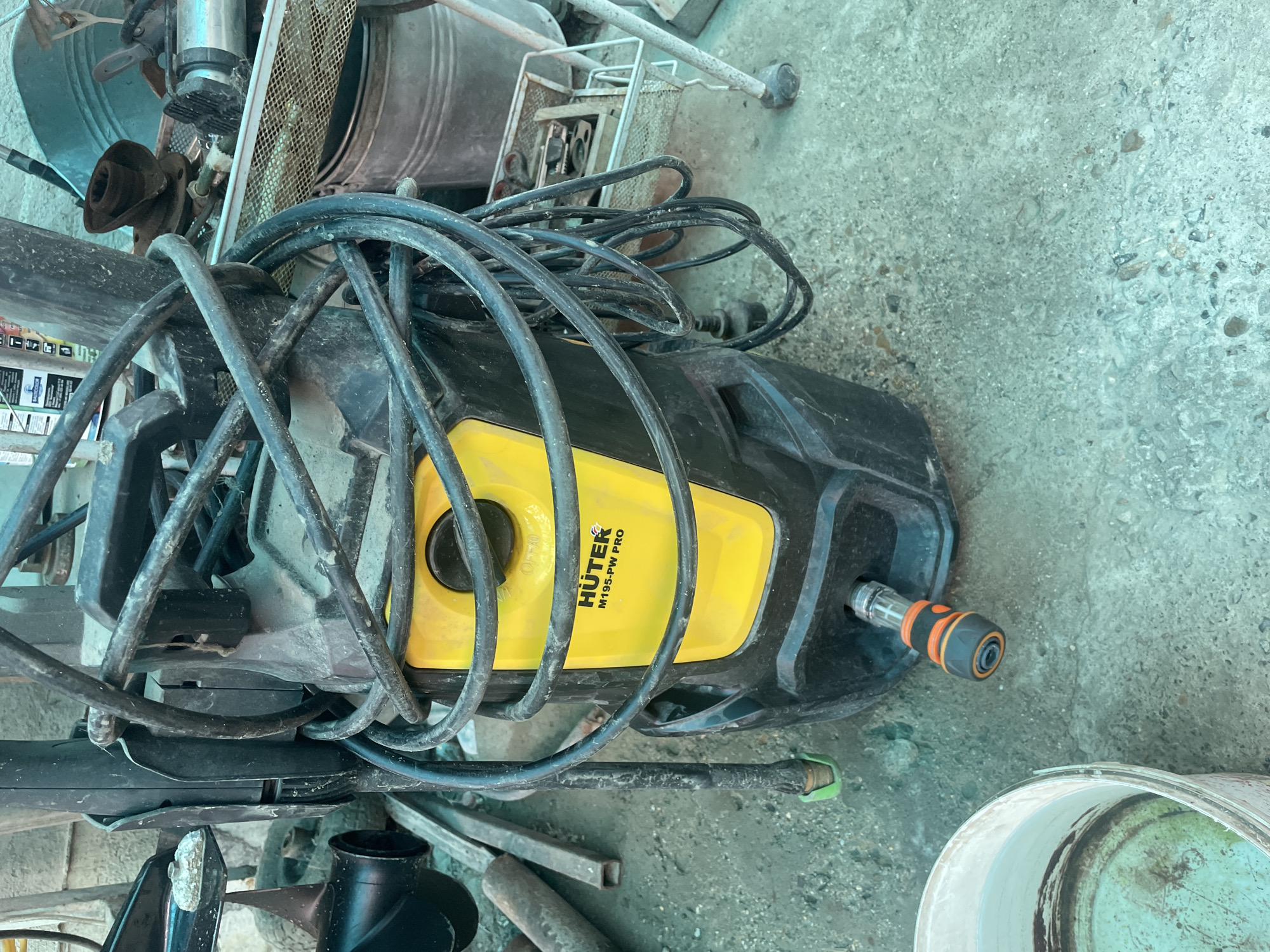 The image shows a Huter M195-PW Pro pressure washer on a concrete surface. The pressure washer is black and yellow, with a coiled black hose draped over it. A hose attachment is visible on the side. Other objects in the scene include a bucket, metal containers, metal grate, and tools. The location is likely a storage area or garage, but it is not possible to confirm the location as Olginka, Russia, based solely on this image.