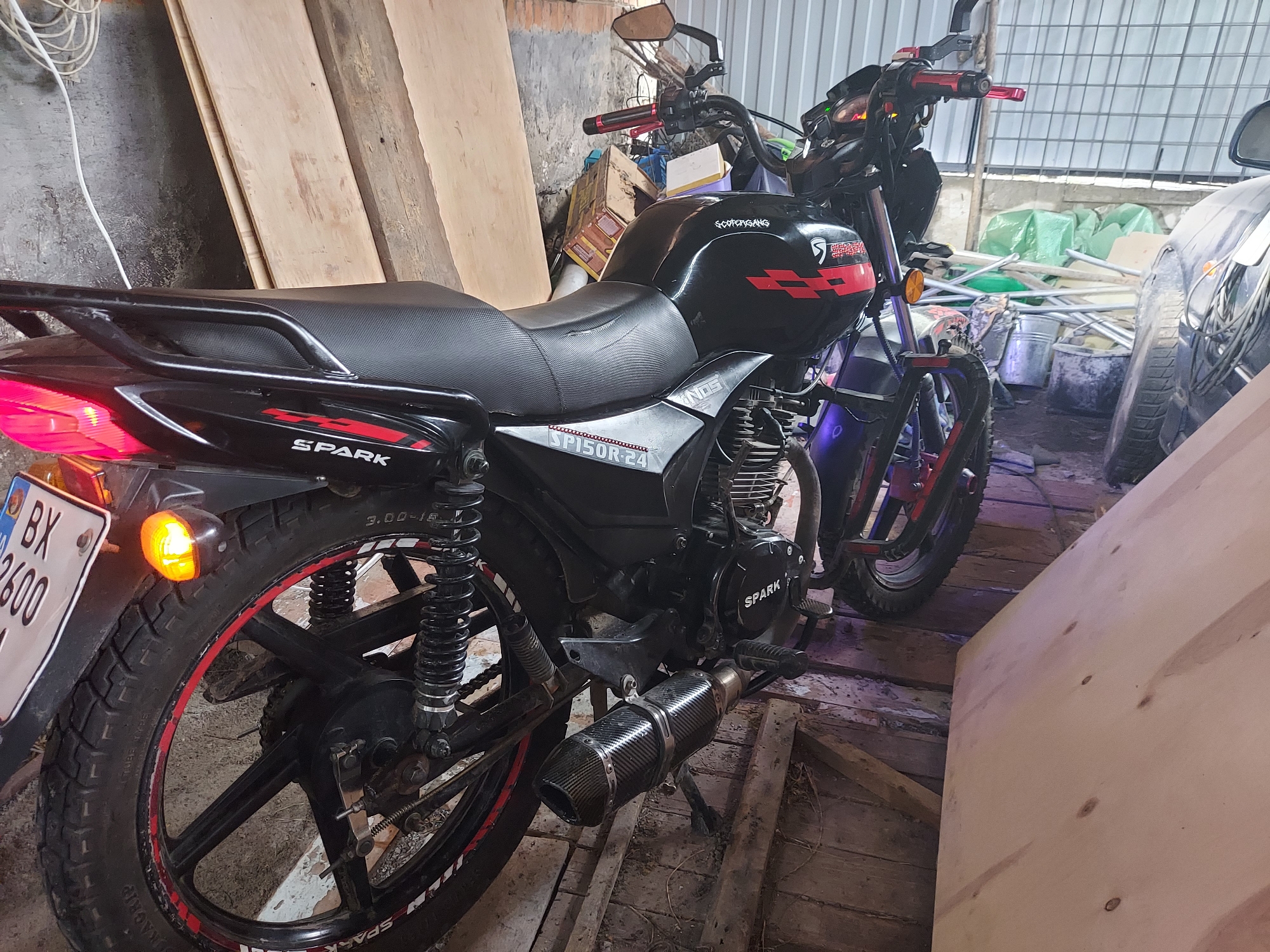 A black Spark SP150R-24 motorcycle is positioned diagonally in an indoor, cluttered storage or garage environment in Unknown, Unknown. The motorcycle, facing the viewer's left, features a black frame, fuel tank, and seat with red graphic accents and "SPARK" branding. The fuel tank displays a "SCOTCHGANG" sticker and a red "S" logo. The seat has a textured, possibly carbon-fiber-patterned material. An aftermarket carbon-fiber patterned exhaust pipe is mounted on the left side. The black alloy wheels have red rim stripes and "SPARK" lettering. The rear tire shows "3.00-18" markings, and the left rear turn signal is illuminated amber. A partially visible license plate begins with "BX 2600". The engine casing is black with a "SPARK" logo. The handlebars are equipped with red levers and grips. The motorcycle rests on its kickstand. The surrounding environment consists of unfinished wooden plank walls on the left, with stacked plywood sheets and cardboard boxes. The floor is composed of uneven wooden planks. In the background on the right, various items including green tarps, metal poles, buckets, and the front wheel and fender of a light-colored vehicle are visible. The ceiling structure appears to be metal. No individuals are present in the scene.