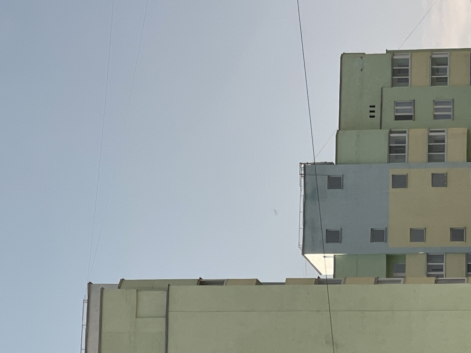 The image shows a partial view of a tall building in Chaikovsky, Russia, composed of several sections with different colors: light green, blue, and yellow. The building has numerous windows. Two thin wires run across the frame from top to bottom. The sky is light blue and clear.