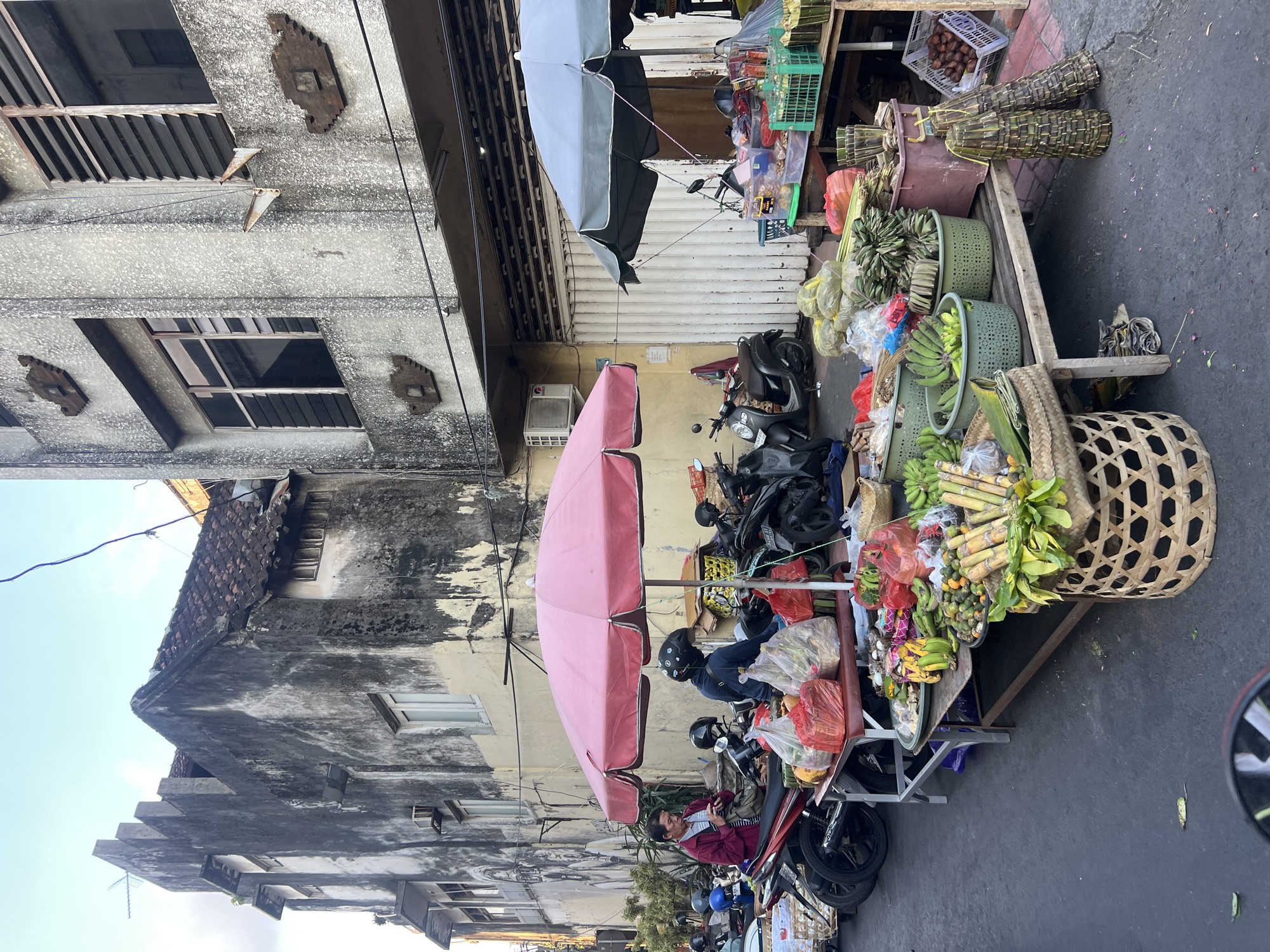 The image depicts a street scene in Denpasar, Indonesia, likely a market or roadside stall. In the foreground, a vendor is attending to a display of fruits and vegetables arranged on tables and in baskets. Items include bananas, sugarcane, and what appear to be coconuts or similar round fruits. Several motorcycles are parked behind the stalls, suggesting a busy urban environment. Two individuals are visible, one in a red jacket interacting with a smartphone, and another wearing a helmet and blue jacket, possibly a customer or vendor. The background shows a weathered, multi-story building with an aged facade, typical of older architecture in the region. A large pink umbrella shades part of the fruit stand, and a smaller green umbrella is visible further down the street. The ground is paved asphalt, with scattered leaves and debris. The overall impression is one of a vibrant, everyday commercial activity on a city street.