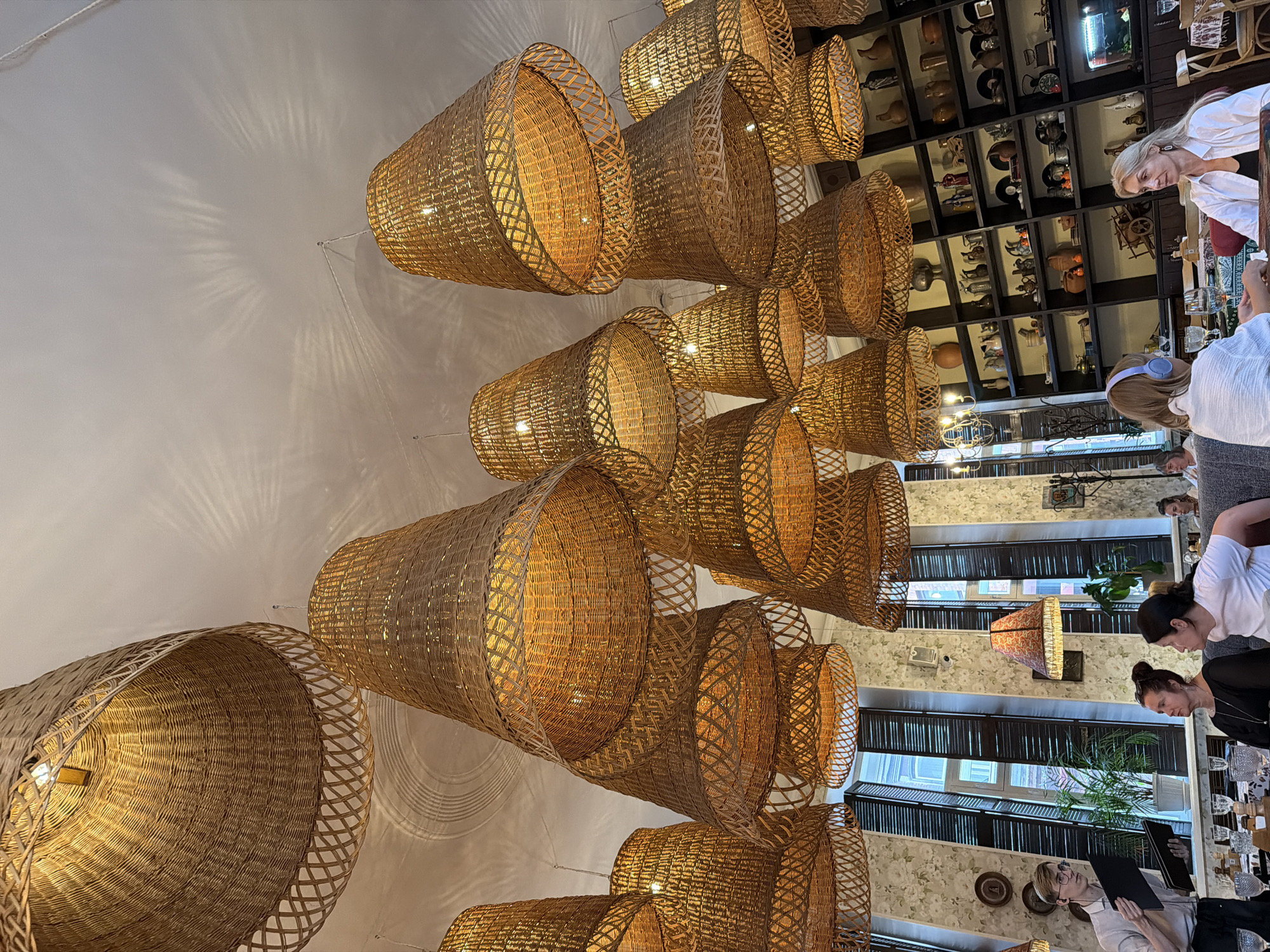 This image depicts the interior of a restaurant or cafe, likely in Krasnoyarsk, Russia. The scene is characterized by an abundance of large, woven basket-like pendant light fixtures, creating a warm and textured ambiance. The lights cast intricate shadows on the white ceiling, suggesting it is daytime or that the interior lighting is significant.
Several people are visible in the image, mostly seen from the back or side as they are seated at tables. One woman with blonde hair, wearing a white shirt, is looking down, possibly at a menu or an object on the table. Another woman with dark hair and headphones is also looking downwards. Further down, another individual is wearing glasses and holding a menu. The presence of menus and table settings indicates that patrons are dining or preparing to dine.
In the background, shelves are filled with various decorative objects, including what appear to be pottery, bottles, and other trinkets, suggesting a rustic or eclectic decor style. There are also large wooden shutters on the windows, adding to the overall aesthetic. The general atmosphere appears relaxed and casual, with no specific event being overtly depicted. The focus is on the unique lighting and the dining setting.