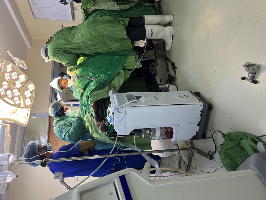 An operating theater in a hospital located in Nairobi, Kenya, identified by "KEMSA" branding on rubber boots, depicts an ongoing surgical procedure. At least five individuals are present: one patient and four medical personnel. The patient lies supine on a metal operating table, largely obscured by green surgical drapes, with a dark-skinned hand visible on the left. Four medical professionals, wearing surgical scrubs (blue and green), caps, and face masks, surround the table.
On the far left, a person in blue scrubs and a blue surgical cap stands observing, positioned near an IV pole from which multiple clear fluid bags are suspended. In the center, a person in green scrubs and a green surgical cap leans over the patient, actively engaged in the procedure. Behind the patient's head, another individual wearing green scrubs and a white head covering is partially visible. On the right, a fourth person in green scrubs and a green surgical cap is actively working on the patient, wearing distinctive white rubber boots with "KEMSA" printed on them.
Overhead, a large, multi-faceted surgical light fixture illuminates the operating field. In the foreground, a white, wheeled medical machine, possibly an anesthesia unit or surgical suction, stands with a transparent collection jar containing a reddish-brown fluid, likely blood. The base of this machine shows signs of wear and minor damage. Below it, a crumpled green cloth rests on the light-colored floor, and to its right, a metal clamp lies detached on the floor. A whiteboard with faint writing is mounted on the wall to the far right. The environment features light-colored walls and flooring, characteristic of a sterile medical facility.