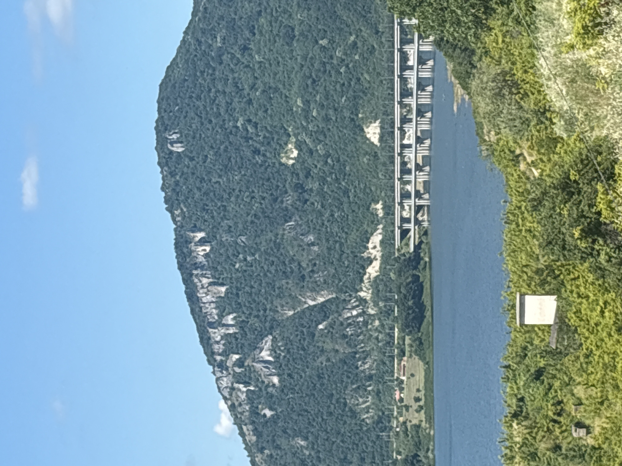 A wide shot captures a serene landscape under a clear blue sky. In the foreground, lush green foliage creates a textured foreground. A simple, white rectangular structure, possibly a small building or marker, is partially obscured by the greenery. Beyond the foliage, a calm blue body of water stretches across the mid-ground. A long, multi-arched bridge spans the water, connecting the two sides. In the background, a massive, tree-covered mountain dominates the scene, with stark white rocky outcrops visible on its slopes. The mountain's peak is rounded and slopes gently downwards. The overall impression is one of natural beauty and tranquil waters.