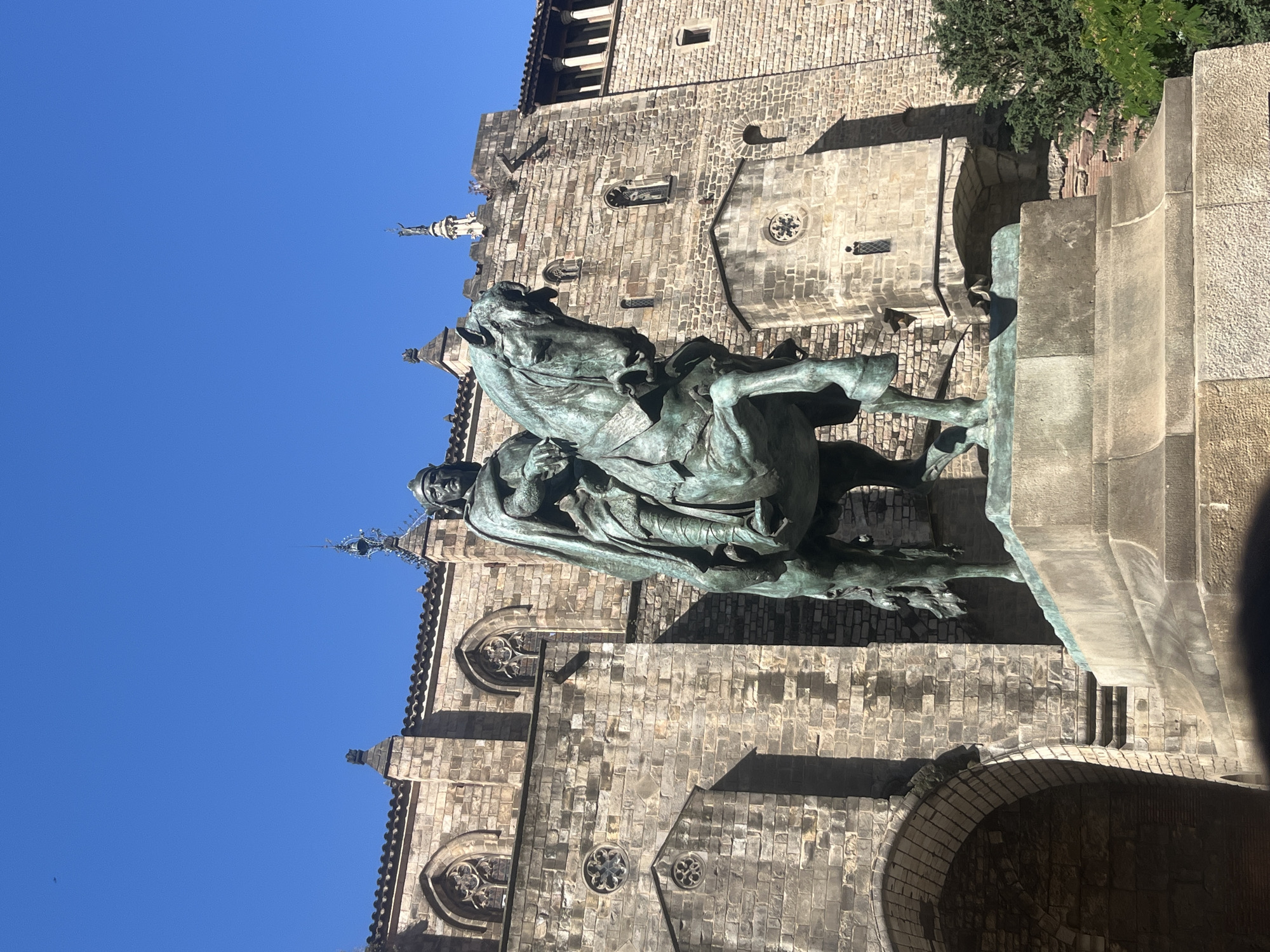 The image captures the equestrian statue of Ramon Berenguer III, Count of Barcelona, standing proudly against the backdrop of the Chapel of Saint Agatha and the Palau Reial Major in Barcelona's Gothic Quarter, Spain. The statue, rendered in bronze, features the count atop a rearing horse, exuding a sense of power and authority. The architectural details of the chapel and palace, characterized by weathered stone walls, arched windows, and pointed spires, contribute to the historical ambiance of the scene. The statue stands upon a stone plinth. The sky is blue.