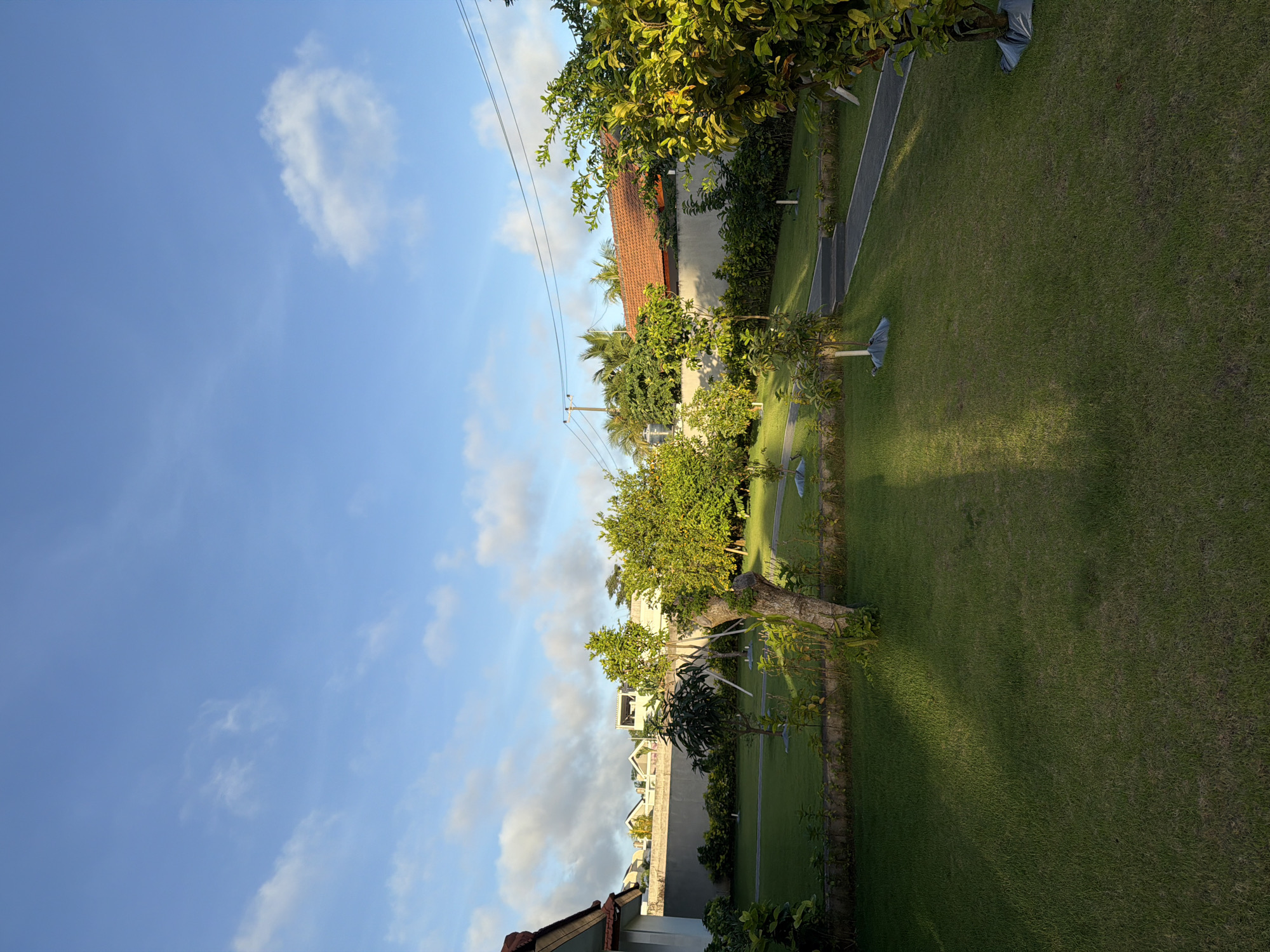 The image depicts a residential area in Kuta Selatan, Indonesia, during daylight. The sky is blue with scattered clouds. The foreground shows a well-maintained lawn bordered by trees and shrubs. Buildings with tile roofs are visible in the background. Utility poles and power lines cross the sky. There are no people visible.
