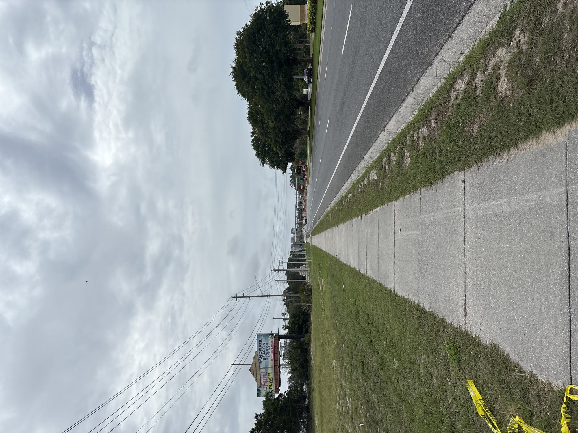 The image shows an outdoor scene in Panama City Beach, Florida, USA. It's taken from the sidewalk alongside a road, looking towards the distance. A multi-lane road is visible with a solid white line dividing the lanes. The sidewalk is concrete with patches of grass on either side. Utility poles and power lines run along the left side of the road. A billboard for "Sandpiper Beacon Beach Resort GulfFront" is visible on the left. The sky is overcast with gray clouds. There is yellow caution tape in the foreground on the right. There are some sparse trees in the horizon. There are no people visible in the image.