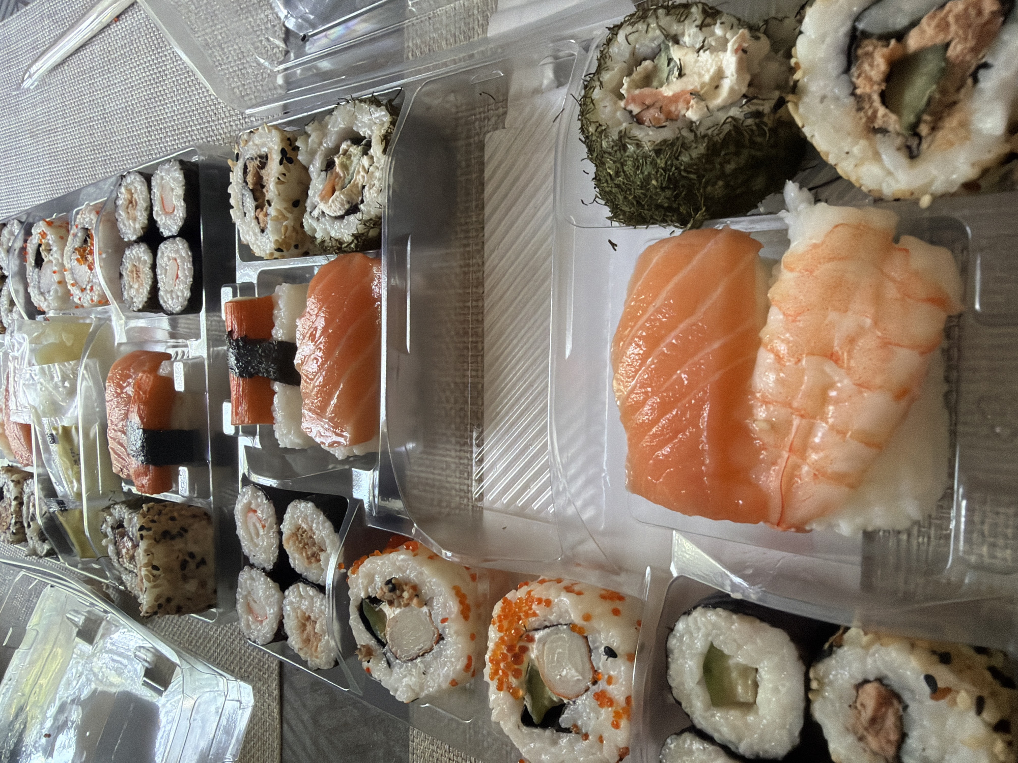 The image displays a close-up, overhead view of a large assortment of sushi and sashimi arranged in clear plastic containers. The food is presented in multiple rows, suggesting a take-out or catering order.
In the top section of the image, several pieces of nigiri sushi are visible, characterized by a slice of fish atop a bed of rice. Some of these are topped with what appears to be crab or imitation crab. There are also some maki rolls with sesame seeds on the outside.
Moving down, there are more maki rolls with various fillings, some featuring orange tobiko (flying fish roe). Further down, there are pieces of salmon sashimi, and a couple of rolls covered in green herbs. In the bottom portion of the image, more maki rolls with visible fillings of tuna or salmon and cucumber are present. There are also two pieces of nigiri, one with salmon and the other with shrimp, placed on rice.
The sushi is arranged on a woven placemat, and a white plastic disposable chopstick is visible on the right side of the image. The overall lighting suggests an indoor setting, likely a home or a casual dining environment. There are no people visible in the image.
The specific location of the city of Sofia, Bulgaria, is not determinable from the visual information provided in the image.
