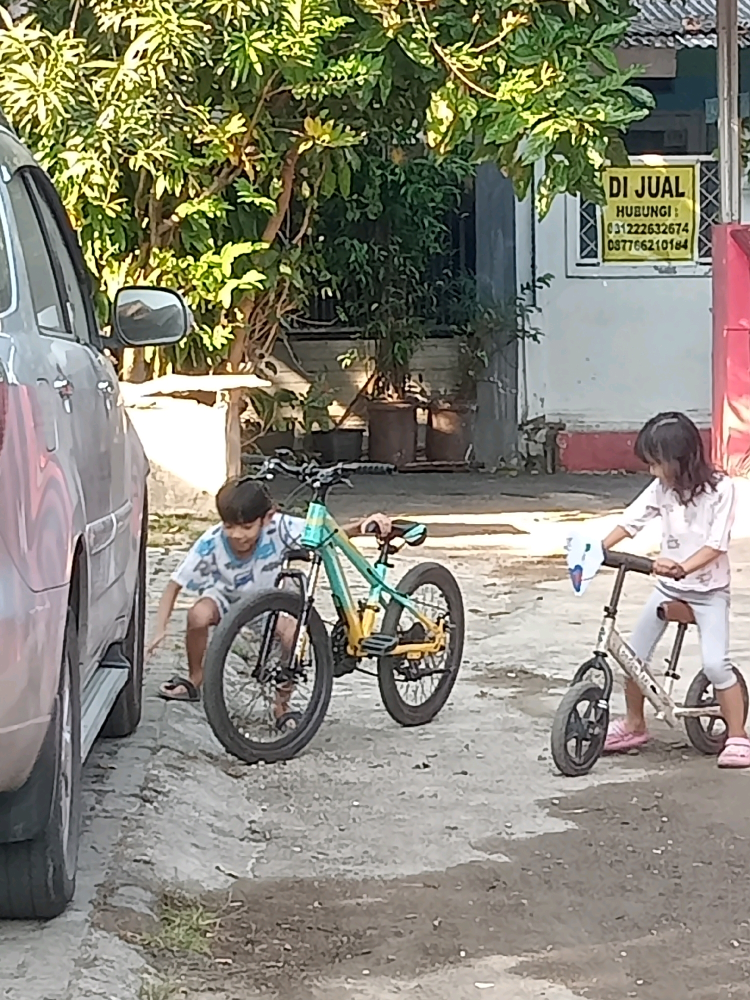 In Cirebon, Indonesia, two children are near bicycles on a paved area. A boy in a patterned shirt and shorts kneels beside a green and yellow bicycle, appearing to interact with it. To the right, a girl wearing a light-colored shirt and pants sits on a smaller bicycle, holding onto the handlebars. A silver car is partially visible on the left, parked next to the children. The background features lush green trees, a building with a "DI JUAL" (For Sale) sign, and what appear to be large planters or barrels. The scene is illuminated by daylight, suggesting an outdoor setting.