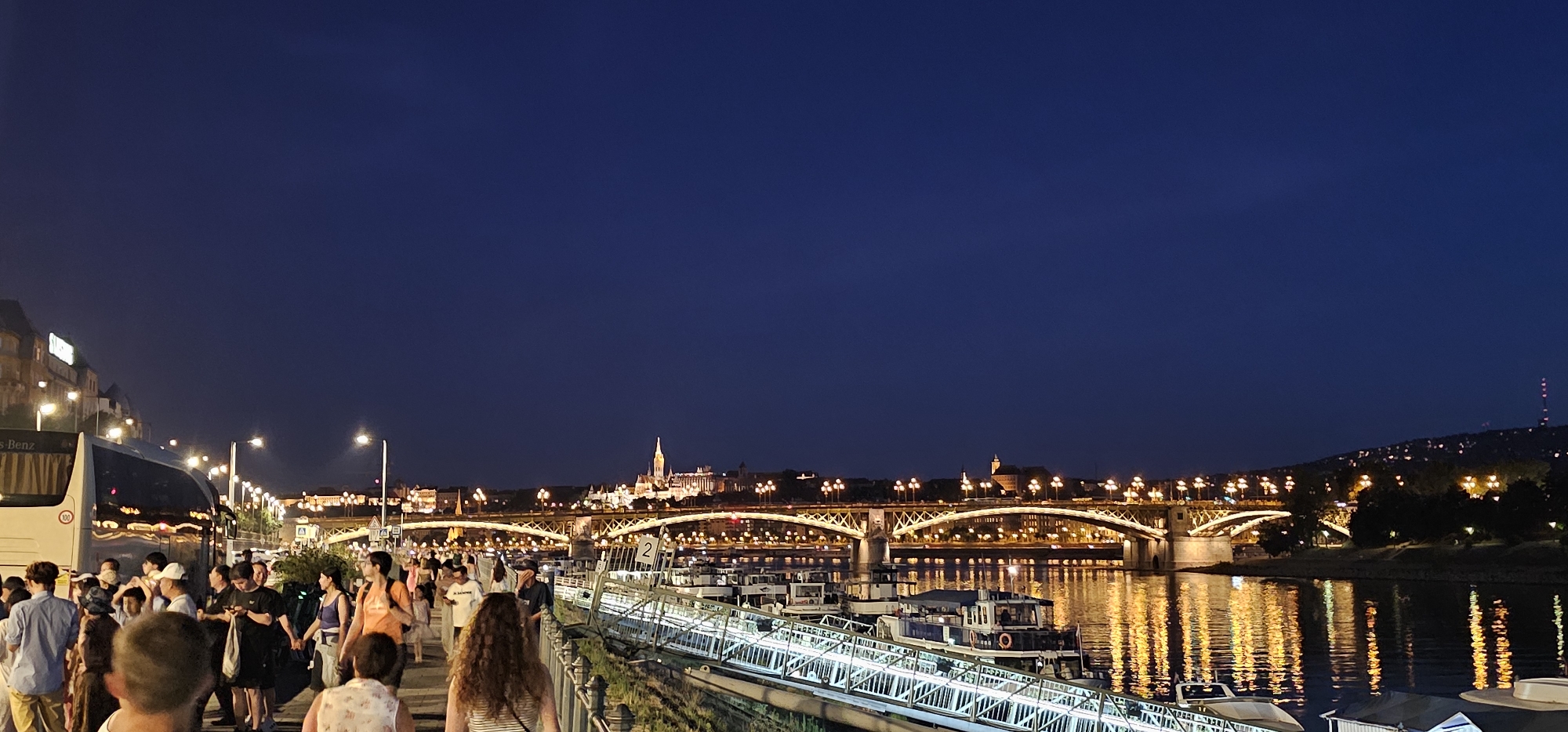 This is a panoramic, horizontal, nighttime, medium-shot photograph taken along the Danube River promenade in Budapest, Hungary. The sky is a deep, clear navy blue. In the background, the Széchenyi Chain Bridge spans the river, illuminated by numerous yellow lights. Beyond the bridge, on the Buda side, the illuminated cityscape of Budapest is visible, including the Buda Castle and Fisherman's Bastion, perched on a hill. The lights of the city and the bridge are reflected in the dark, rippling water of the Danube.
In the foreground, a paved promenade is filled with a diverse crowd of people walking. Many are looking towards the bridge and the illuminated cityscape. The people are casually dressed, suggesting it is a warm evening. To the left, a white tour bus with "Mercedes-Benz" branding is parked. Along the right side of the promenade, a row of docked boats and a white railing border the river. The overall atmosphere is one of evening leisure and sightseeing.