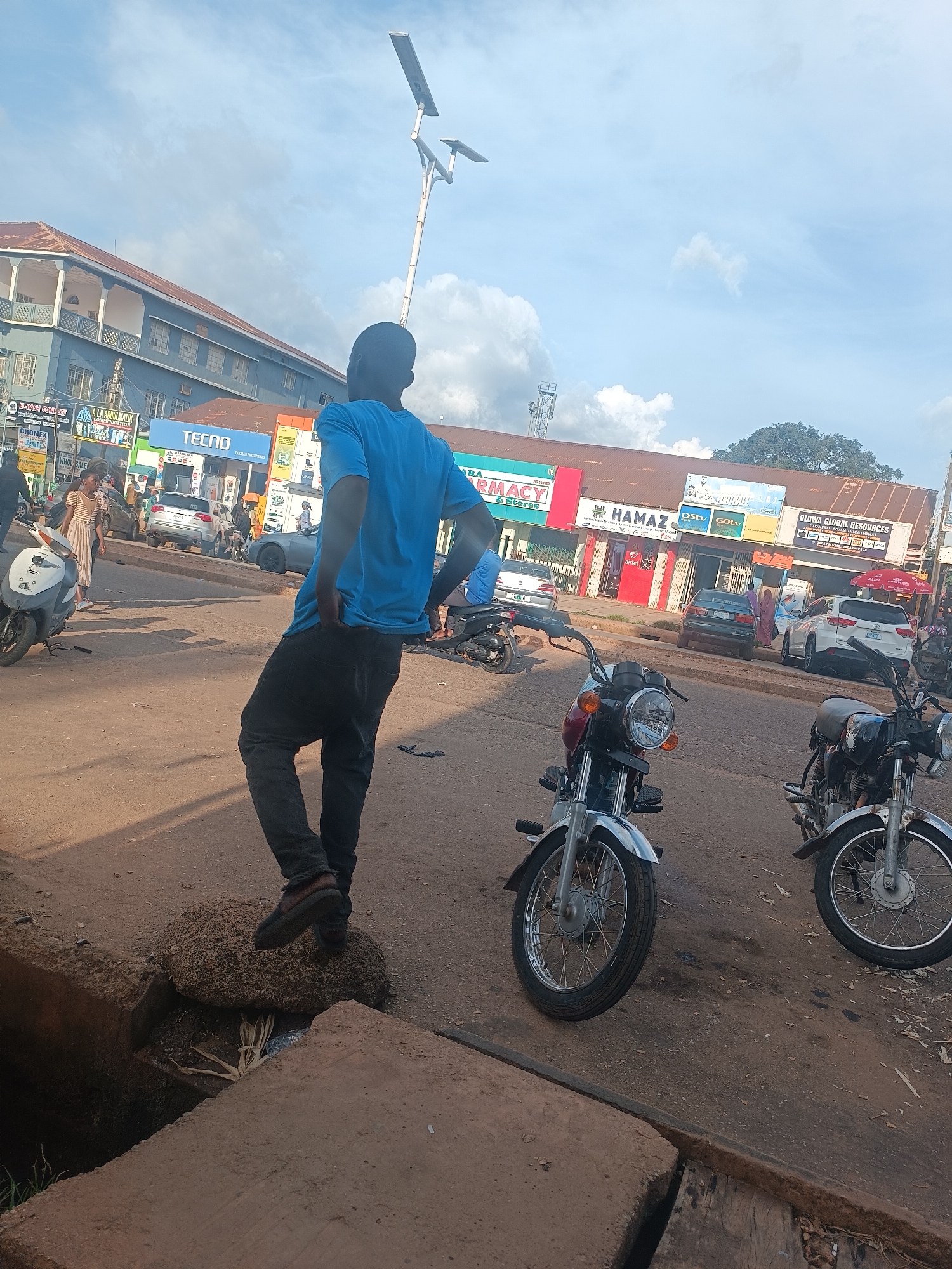 The image captures a street scene in Jos, Nigeria, on a partly cloudy day. A young man wearing a blue t-shirt and black jeans stands facing away from the camera, his hands in his pockets, with his left foot resting on a large rock next to a concrete drainage ditch. In the background, various businesses line the street, including a "TECNO" store, a pharmacy, and "HAMAZ" electronics. Several vehicles, including cars and motorcycles, are parked or driving on the road. A white scooter is visible on the left side of the frame, and two motorcycles are parked in the foreground. The overall impression is that of a typical commercial area in an urban setting.