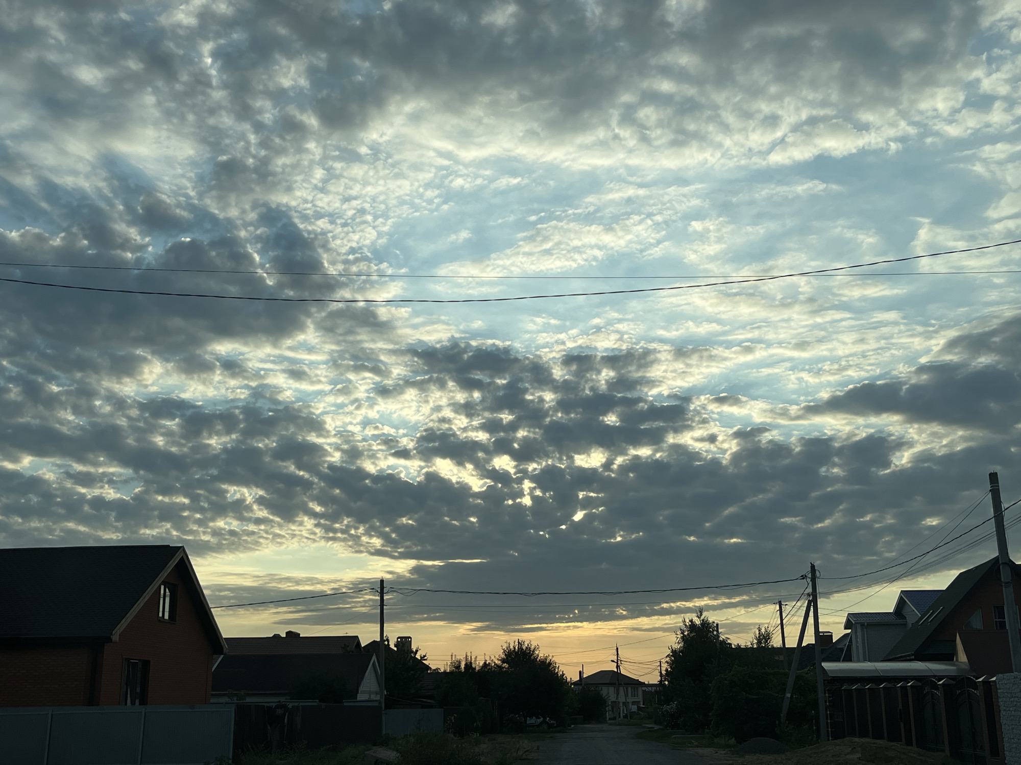 The image depicts a street scene in Taganrog City District, Russia, under a dramatic cloudy sky. The perspective is from ground level, looking down a paved road that curves slightly to the right and disappears into the distance. The sky is filled with a textured layer of cumulus clouds, with patches of brighter sky and sunlight breaking through in places, creating a dappled effect. The sun appears to be low on the horizon, likely during dawn or dusk, casting a warm, yellow-orange glow in the distance.
On either side of the road, residential houses with dark roofs are visible, some with brick facades and windows. Trees and vegetation line the street, creating a sense of a suburban or semi-rural environment. Several utility poles with overhead wires are present, stretching across the scene and contributing to the urban infrastructure. The foreground shows a low fence. No people or animals are discernible in the image. The overall atmosphere is one of quiet observation, with the natural light and weather patterns dominating the visual landscape.
