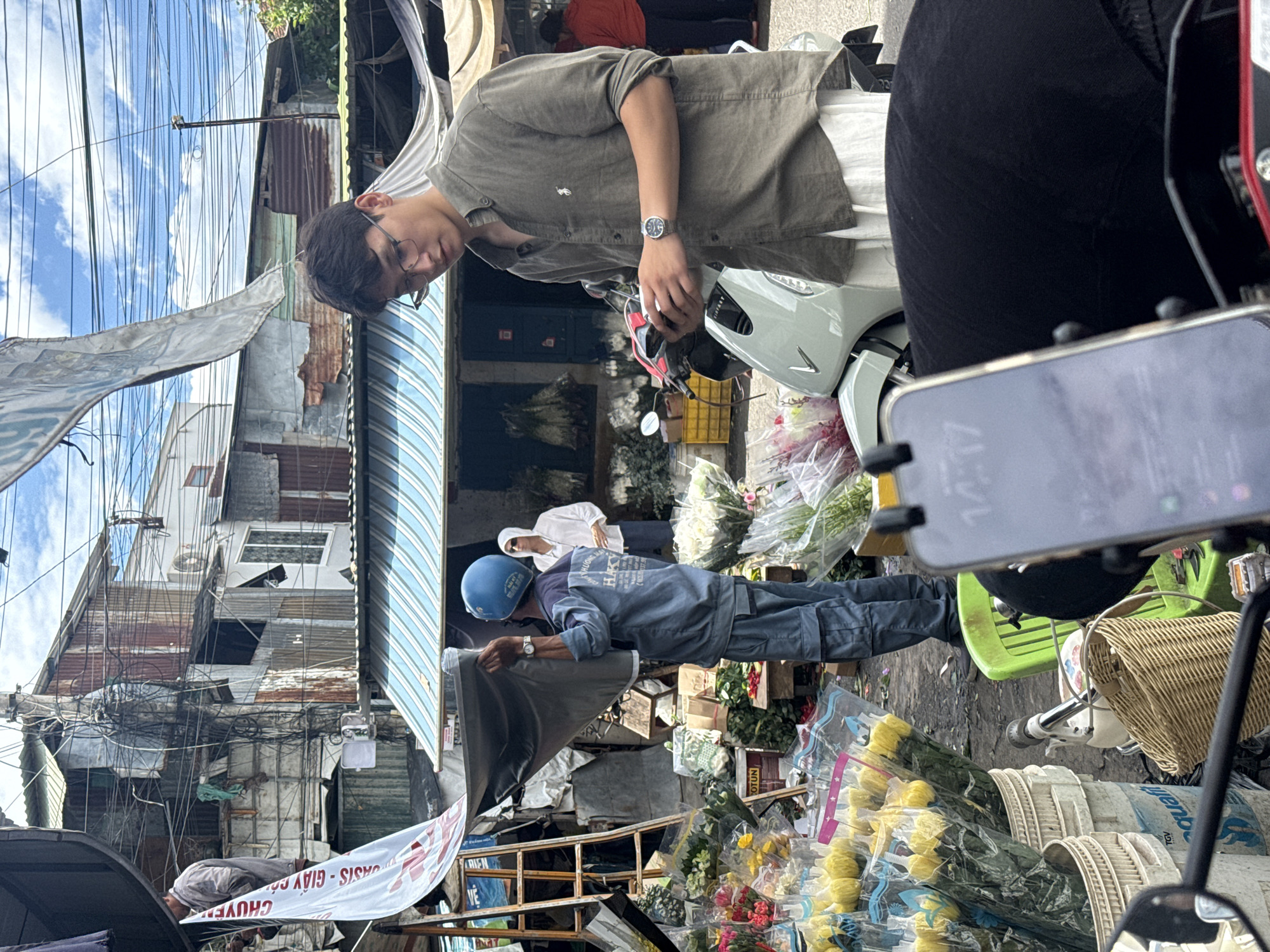 This is a street scene in Nha Trang, Vietnam, likely a flower market given the abundant display of flowers in buckets and plastic wrapping. In the foreground, a person is partially visible, operating a motorcycle, with a smartphone mounted on the handlebars showing the time "14:17". Behind them, a young man wearing glasses and a green button-down shirt stands near a light green scooter, looking at his phone. To his left, a man in a blue uniform and helmet is holding up a large, dark tarp. Behind him, another person in a white hoodie stands near a flower stall. The background shows a chaotic urban environment with dilapidated buildings, numerous electrical wires, and various market stalls covered by awnings.