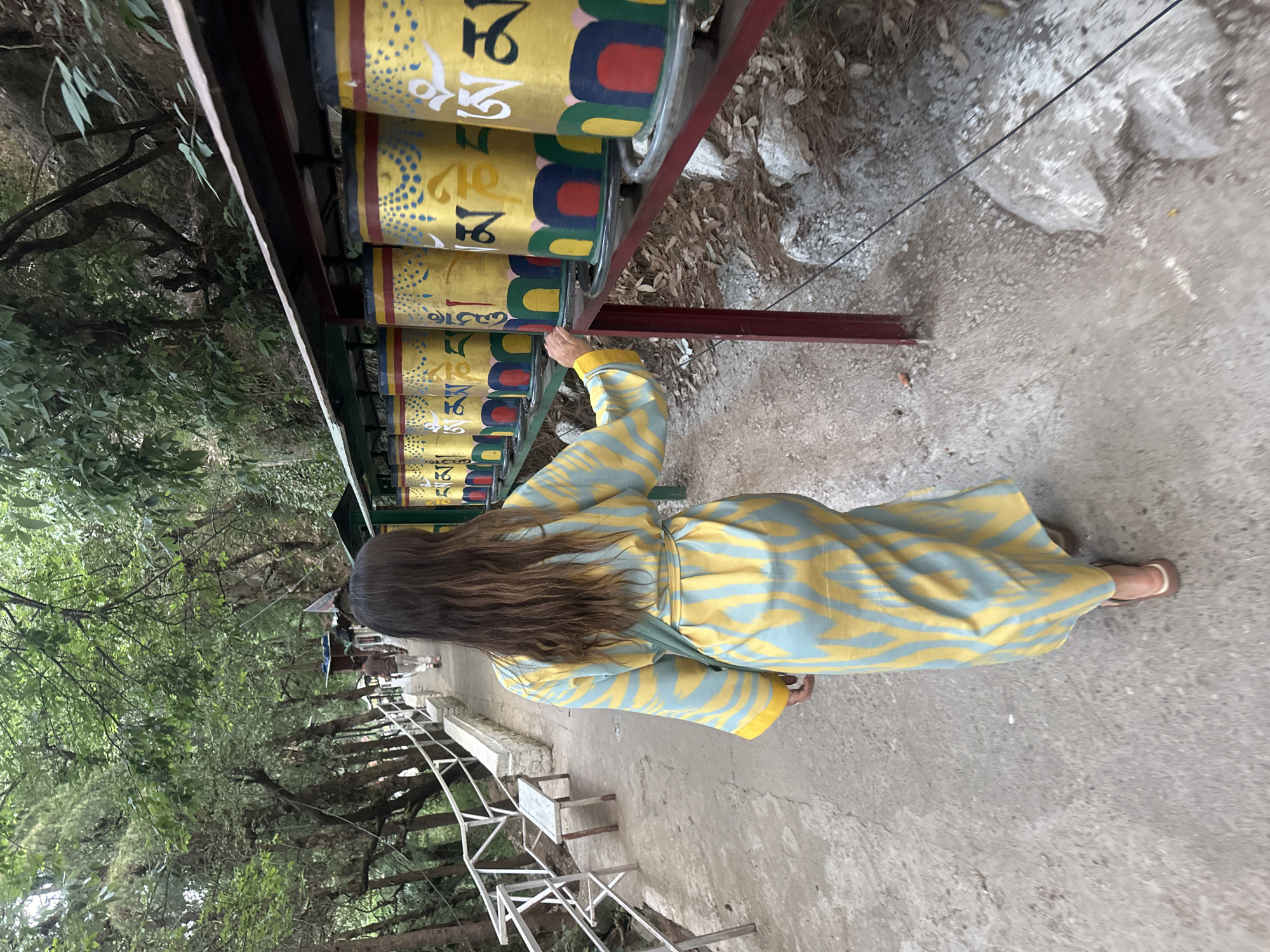 A woman wearing a long, patterned robe is spinning a row of prayer wheels. The prayer wheels are yellow with blue and red accents, and the symbols are written in gold. They are mounted on a metal frame. The location is a paved path in Dharamshala, India. Trees and a metal railing are visible to the left. The ground is stone.