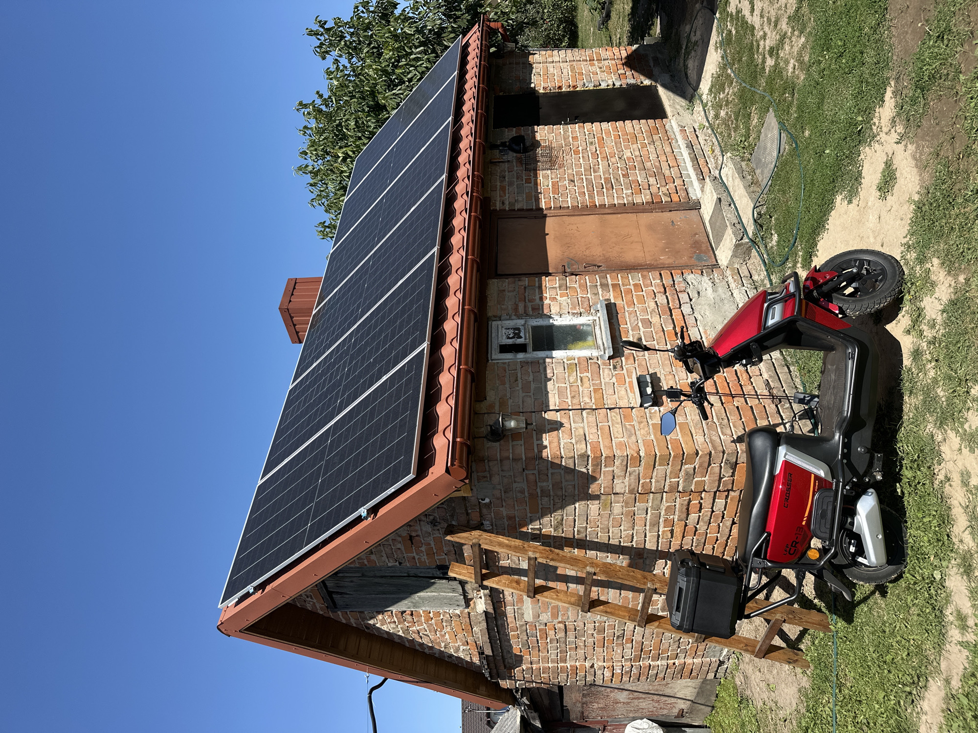 The image depicts a red electric scooter parked on a dirt and grass area in front of a single-story brick building. The building has a sloped roof covered with solar panels, a brown metal chimney, and two brown metal doors. A wooden ladder is propped against the left side of the building. A black storage box is attached to the rear of the scooter. The scene appears to be a residential setting in the village of Zmiinets, Ukraine. The sky is clear and blue.
