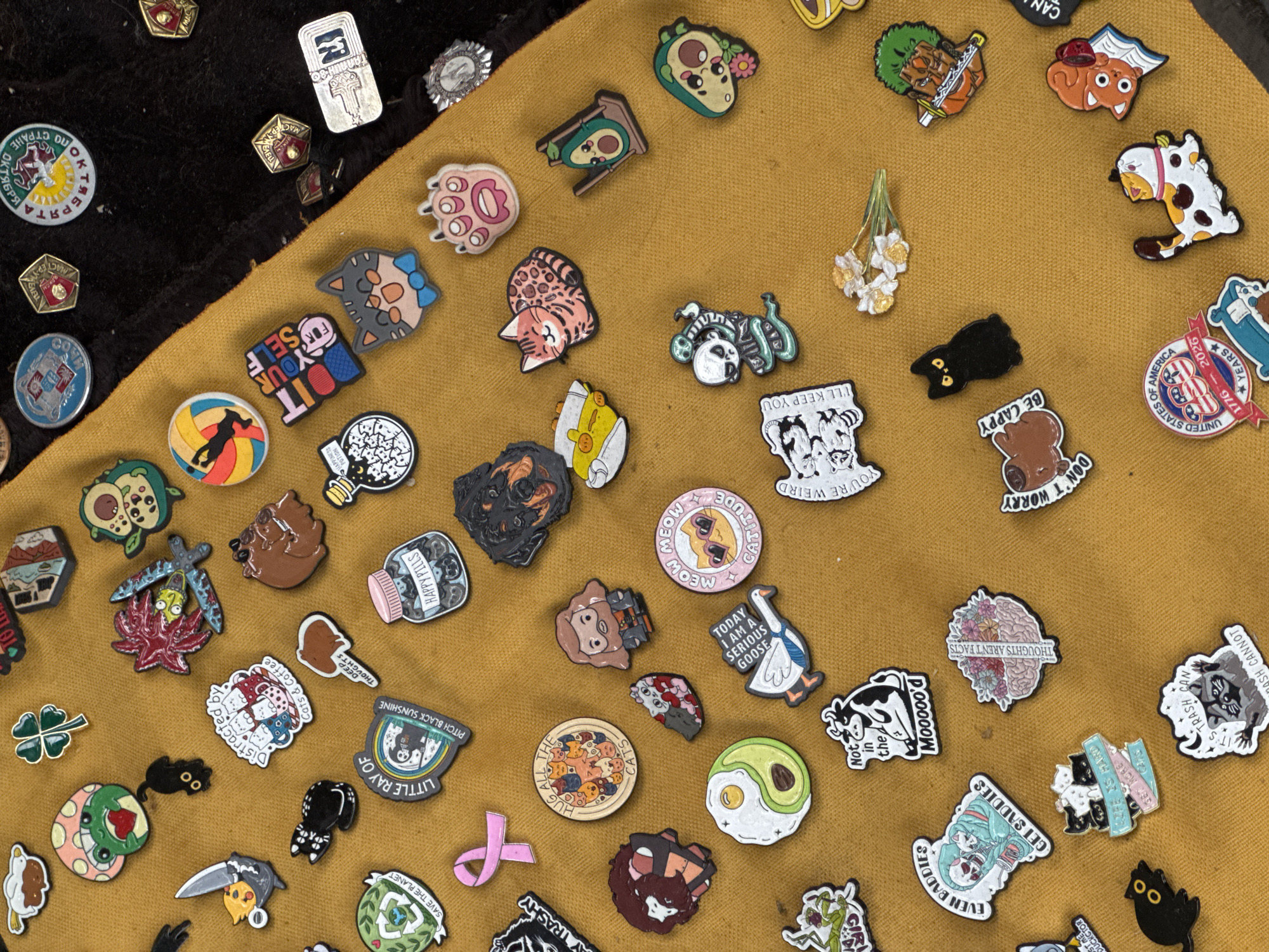 The image displays a collection of enamel pins scattered across a mustard-yellow fabric surface. The pins feature a variety of designs, including animals (cats, dogs, cows, raccoons, frogs), food items (avocado, fried egg), objects (bottles, swords, mushrooms), and text-based messages. Some of the pins have a cartoonish or whimsical aesthetic, while others are more detailed. The pins are not arranged in any particular order, and their placement suggests they have been casually laid out. There is no discernible context for the location, nor is there any information that would allow for its identification as Tbilisi, Georgia. The scene is static and contains no people or actions.