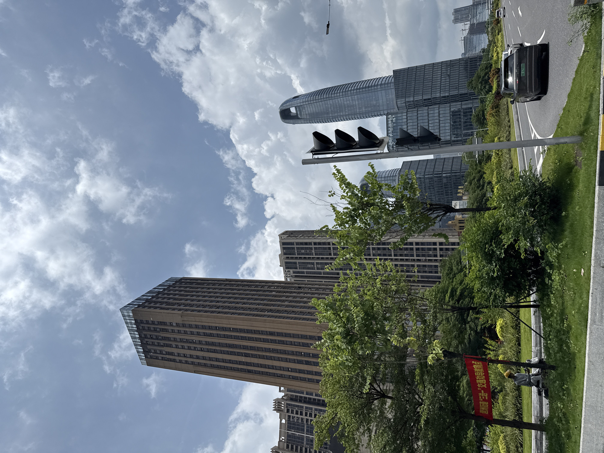 In the Haizhu District of China, on a partly cloudy day, a street scene unfolds with modern architecture and urban elements. Dominating the background are several tall skyscrapers, one of which is a slender, tan-colored building with a glass-topped peak. To its right, a distinctively curved, glass-facade skyscraper stands out. In the mid-ground, a traffic light with three black lights faces the street. A dark SUV with license plate "AA L25666" is driving on the road in the foreground, followed by another white car. To the left of the road, a person wearing a yellow hard hat and a light-colored shirt is seated next to a scooter, with a red banner hanging on a pole nearby. Lush green trees and manicured bushes line the roadside, suggesting a well-maintained urban landscape. The overall impression is one of a bustling, contemporary city environment.