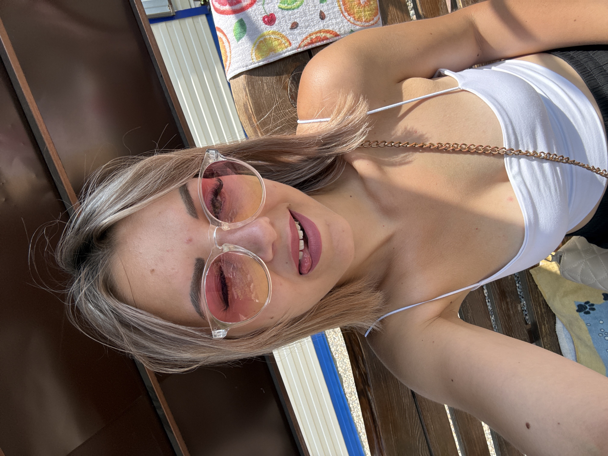A young woman with blonde hair and sunglasses is sitting on a wooden bench outdoors. She is wearing a white tank top and black pants. Her tongue is sticking out, and she is winking. A towel with fruit prints is draped over the bench next to her. The background shows a wooden structure and a striped wall. The scene appears to be in a park or recreational area.