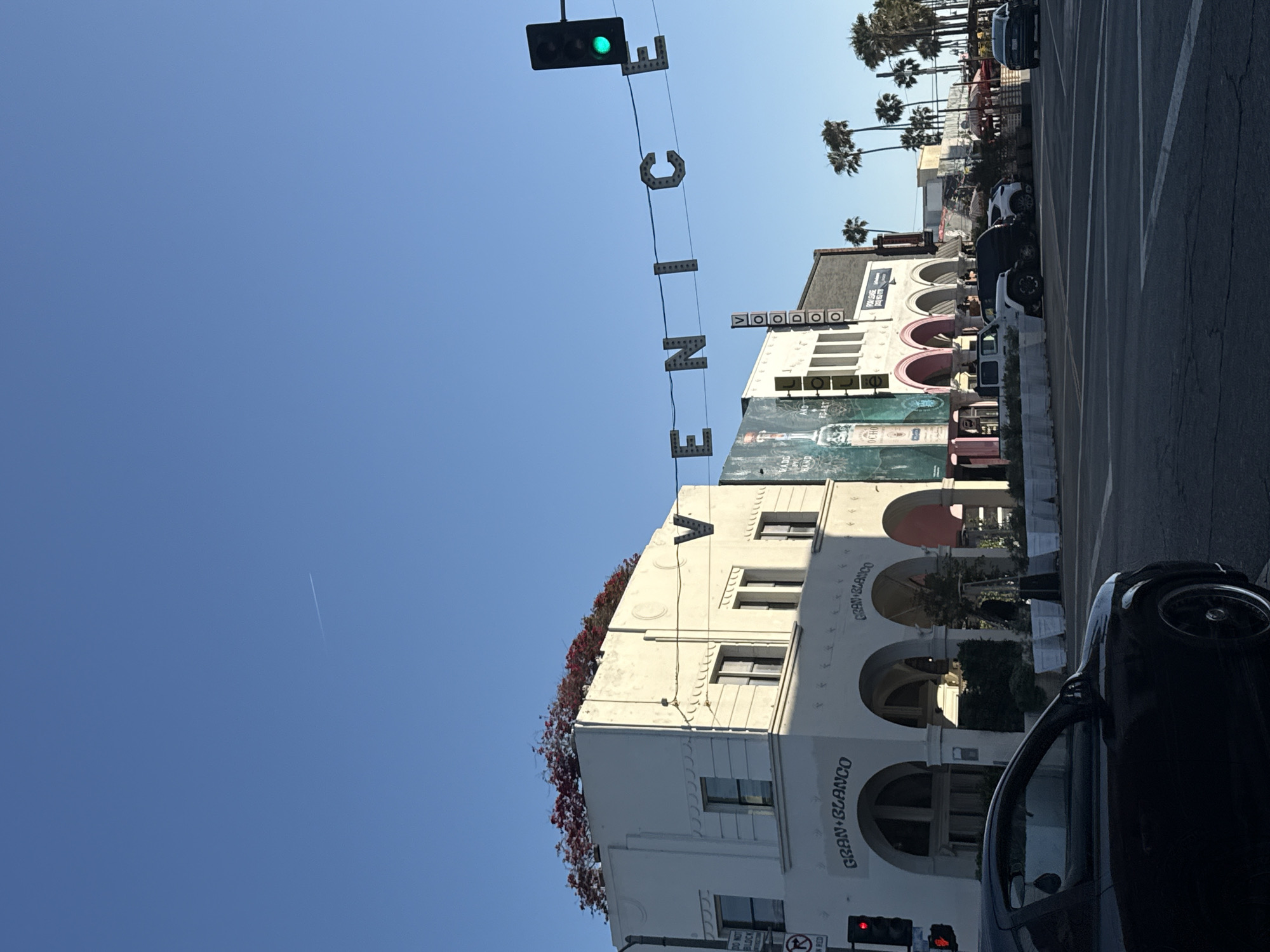 The image shows a street scene in Venice, Los Angeles, United States of America. Overhead, the word "VENICE" is spelled out with individual letter signs strung between a building and a green traffic light. The architecture is predominantly white, Art Deco style, with arched windows and balconies. A building is labeled "GRAN BLANCO." Palm trees are visible in the background. Cars are parked along the street. The sky is clear and blue.