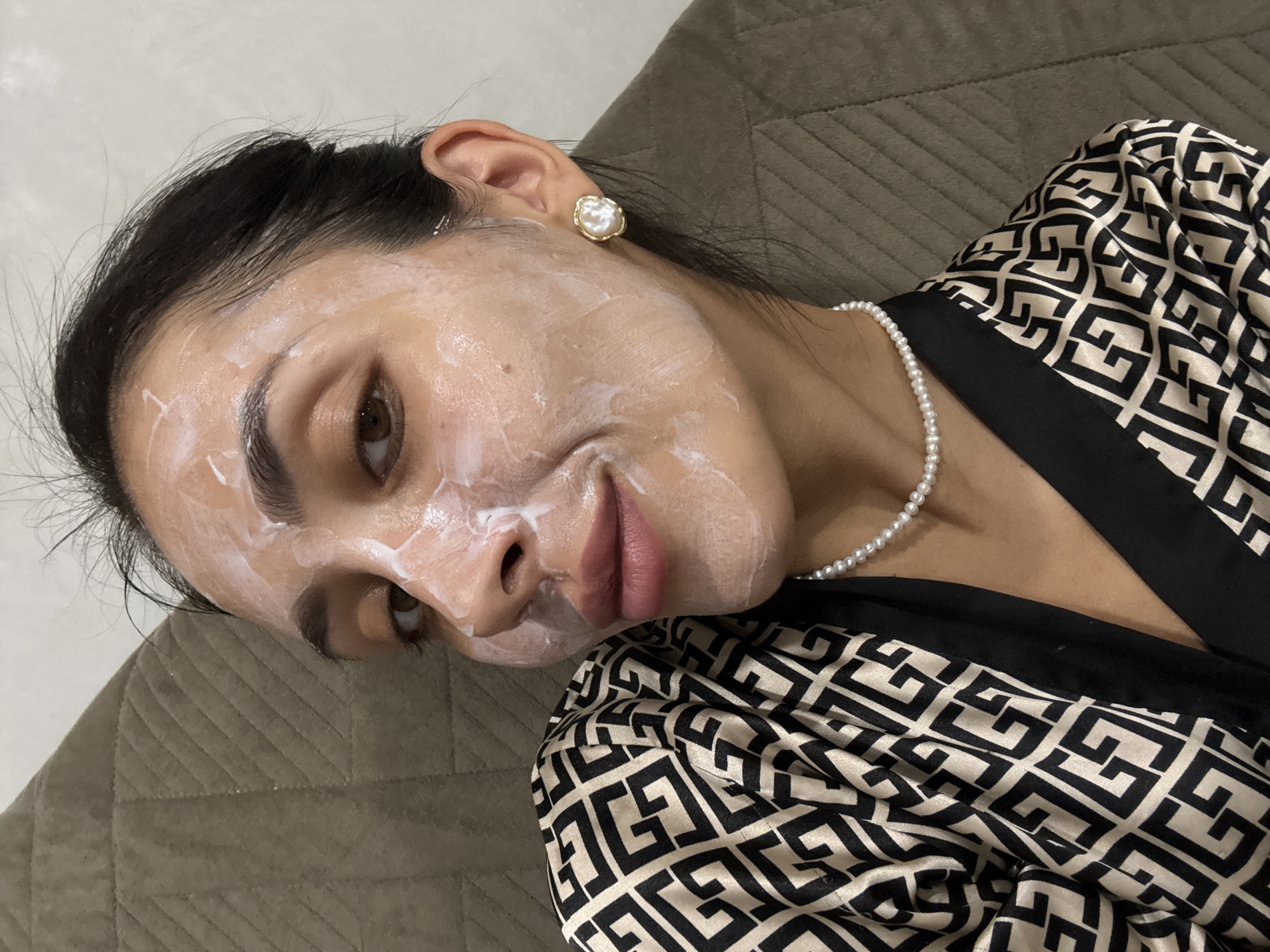 A close-up, high-angle, selfie-style shot captures a woman lying down with a face mask applied to her face. The woman has dark hair, brown eyes, and is wearing a black and beige patterned robe and a pearl necklace. She is also wearing a white and gold earring. The background consists of a textured, brown surface. The image is taken indoors. There is no identifiable information about the location in Votkinsk, Russia.