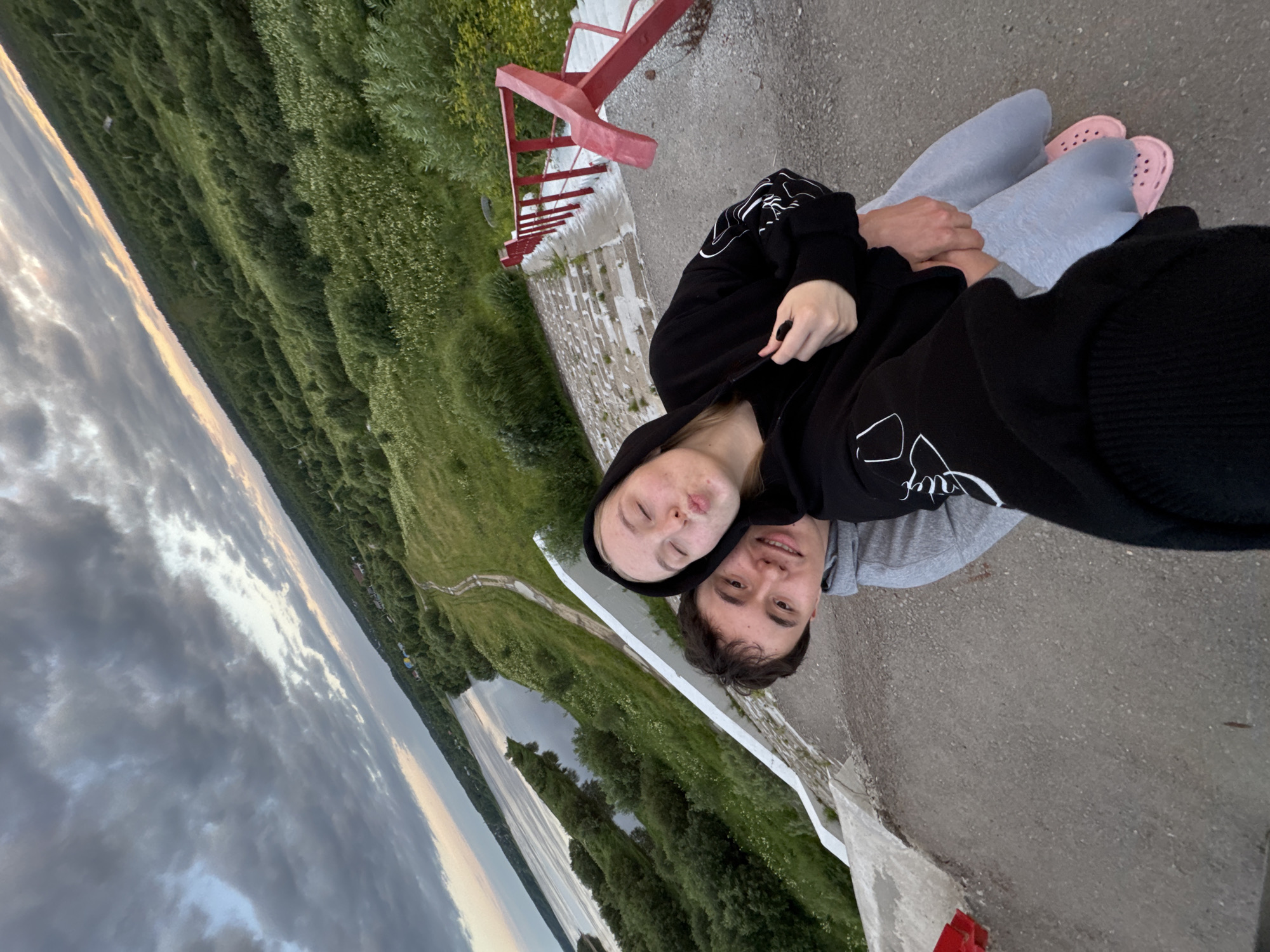 In the city of Ryabinino, Russia, a young couple is taking a selfie outdoors. The man, wearing a grey hoodie, is on the left, with his arm around the woman. The woman, wearing a black hoodie, is on the right, puckering her lips for the photo. They are standing on a concrete surface that appears to be a pathway or staircase. To their right, a red railing runs alongside the stairs. The background features a wide river on the left, with lush green trees and vegetation covering a sloping hill. A dirt path winds through the greenery. The sky above is filled with dramatic, dark clouds, with a sliver of lighter sky visible on the horizon, suggesting it may be dusk or early dawn. The overall scene depicts a natural landscape with a man-made structure.