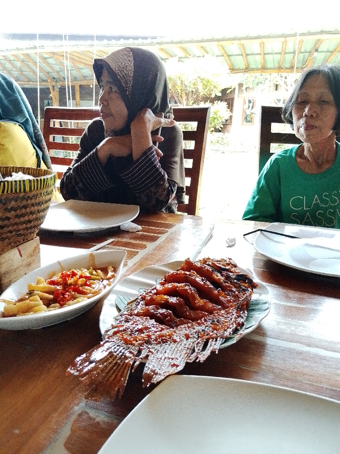 The image depicts an outdoor dining scene in Pabelan, Indonesia, focusing on a wooden table set with food and two women. In the foreground, a large, whole grilled fish, covered in a reddish-brown sauce, rests on a white plate with a banana leaf liner. To its left, a white boat-shaped dish contains fried food, likely tempeh or tofu, mixed with red chili peppers and a sauce. Several empty white plates are also present on the table. On the far left, a woven basket with a light-colored top, possibly containing rice, is visible. Two adult women are seated at the table. The woman on the left, wearing a dark hijab with a patterned band and a dark long-sleeved top, has her right arm resting on the table with her hand near her face, and she is looking off to the right. The woman on the right, wearing a green t-shirt with "CLASSY SASSY" printed in white, is seated facing the viewer with her eyes partially closed. The background reveals an open-air structure with a light-colored roof supported by wooden beams, beyond which green foliage and trees are visible under bright daylight conditions.