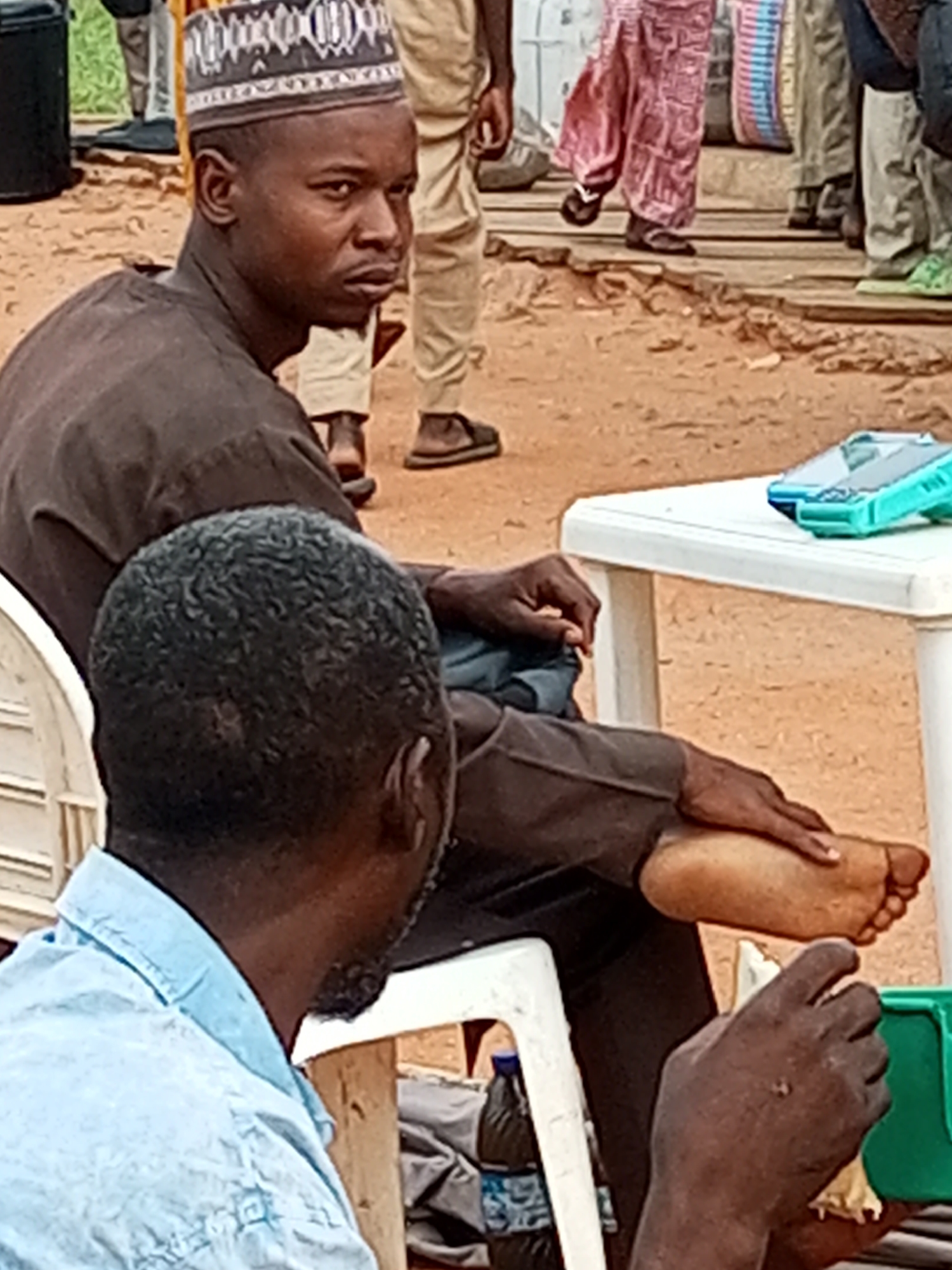 In Gombe, Nigeria, at an outdoor event, two men are visible. The man in the foreground, wearing a light blue shirt, is partially visible from the neck up, with his back to the camera. He has dark, curly hair and is holding something yellow and white in his right hand. To his left and slightly behind him, another man is seated, facing away from the camera, wearing a brown robe and a patterned cap. His legs are crossed, and his right foot, with visible toes, rests on his left thigh. Between his legs is a dark-colored fabric. A white plastic table is to the right of this man, holding two bright turquoise plastic containers. The ground is sandy and uneven, with some scattered debris. In the background, other individuals are blurred and indistinct, with some wearing traditional Nigerian attire.