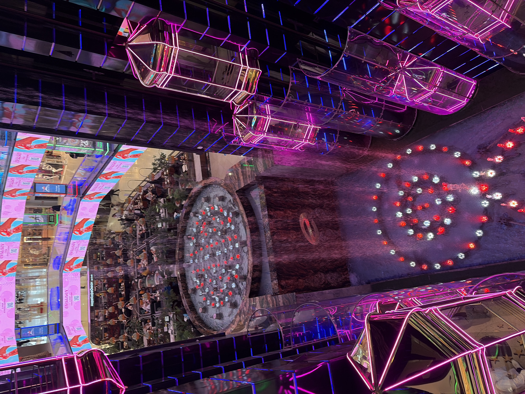 The image depicts the interior of a multi-level shopping mall, likely a modern complex with a focus on elaborate design and entertainment. The scene is dominated by a central atrium featuring a cascading waterfall and a circular fountain with red and white lights at the bottom. Several glass elevators with neon pink lighting are visible, ascending the side of the atrium, reflecting the surrounding lights and architecture. Above, balconies and walkways with rows of cafes and shops are visible, adorned with large digital screens displaying colorful advertisements, primarily featuring vibrant graphics and text in what appears to be Russian. People are sparsely distributed throughout the visible areas, seated at tables in the cafes and walking on the upper levels. The overall atmosphere is one of contemporary retail and entertainment, with a strong emphasis on visual stimulation through lighting and digital displays. The location is identified as Moscow, Russia.