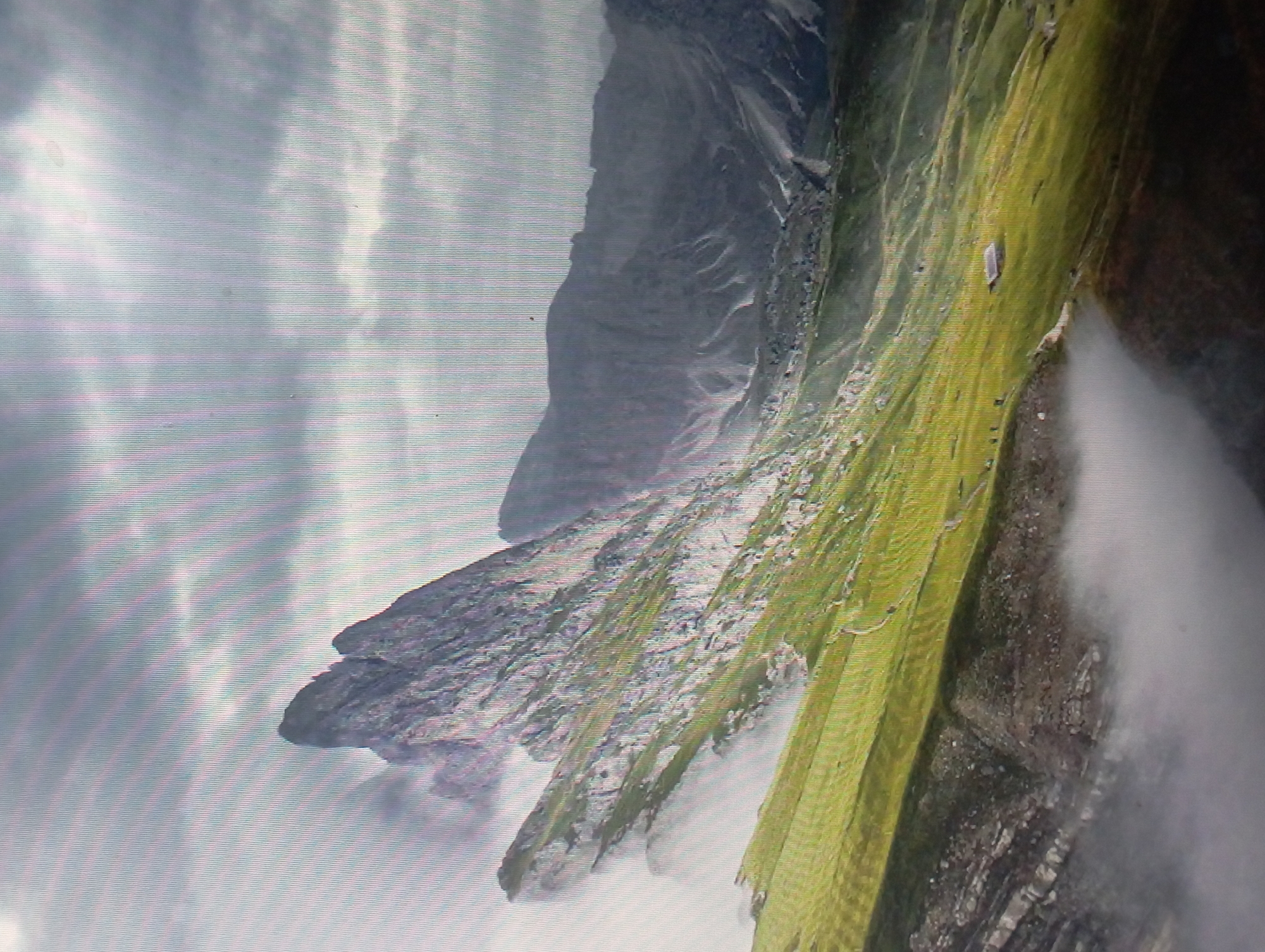 The image shows a mountain landscape, rotated 90 degrees clockwise. The landscape features a tall, rugged mountain on the left side with a dark grey, rocky surface. To the right of the mountain, a steep, grassy slope extends downwards. A waterfall cascades down the right edge of the frame, partially obscured by mist. The sky above the mountain is overcast and cloudy. There is no indication of this photo being taken in the city of Karaganda, Kazakhstan.