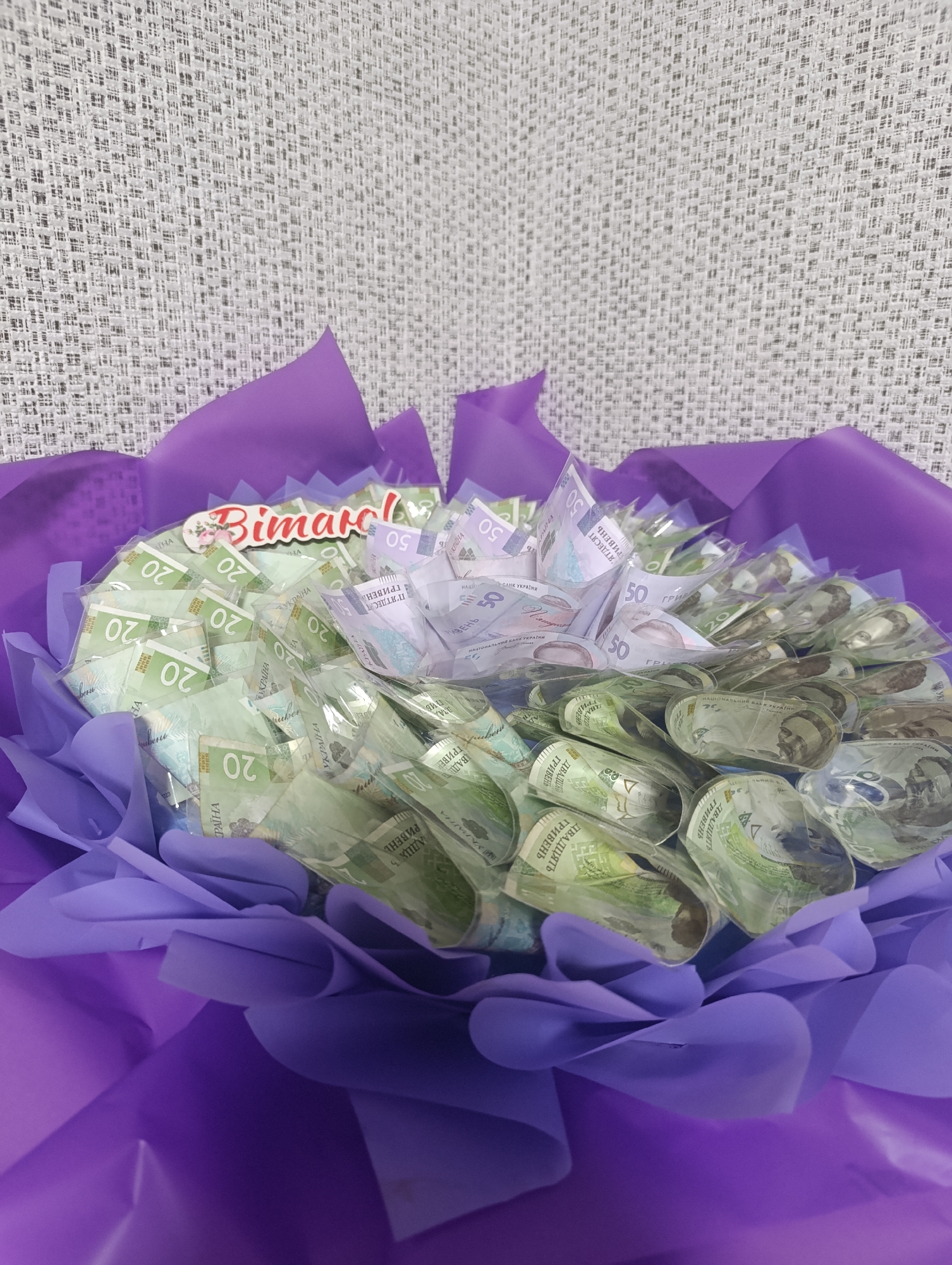 A bouquet of Ukrainian hryvnia banknotes, wrapped in purple paper, is displayed against a textured grey background. The banknotes, predominantly in green and white hues, are rolled and arranged in a circular formation. Some banknotes are clearly visible with denominations of 20 and 50 printed on them. A small decorative element with the word "Bimarol" in red and white lettering, adorned with flowers, is positioned at the top center of the bouquet. The lighting is even, suggesting an indoor setting.