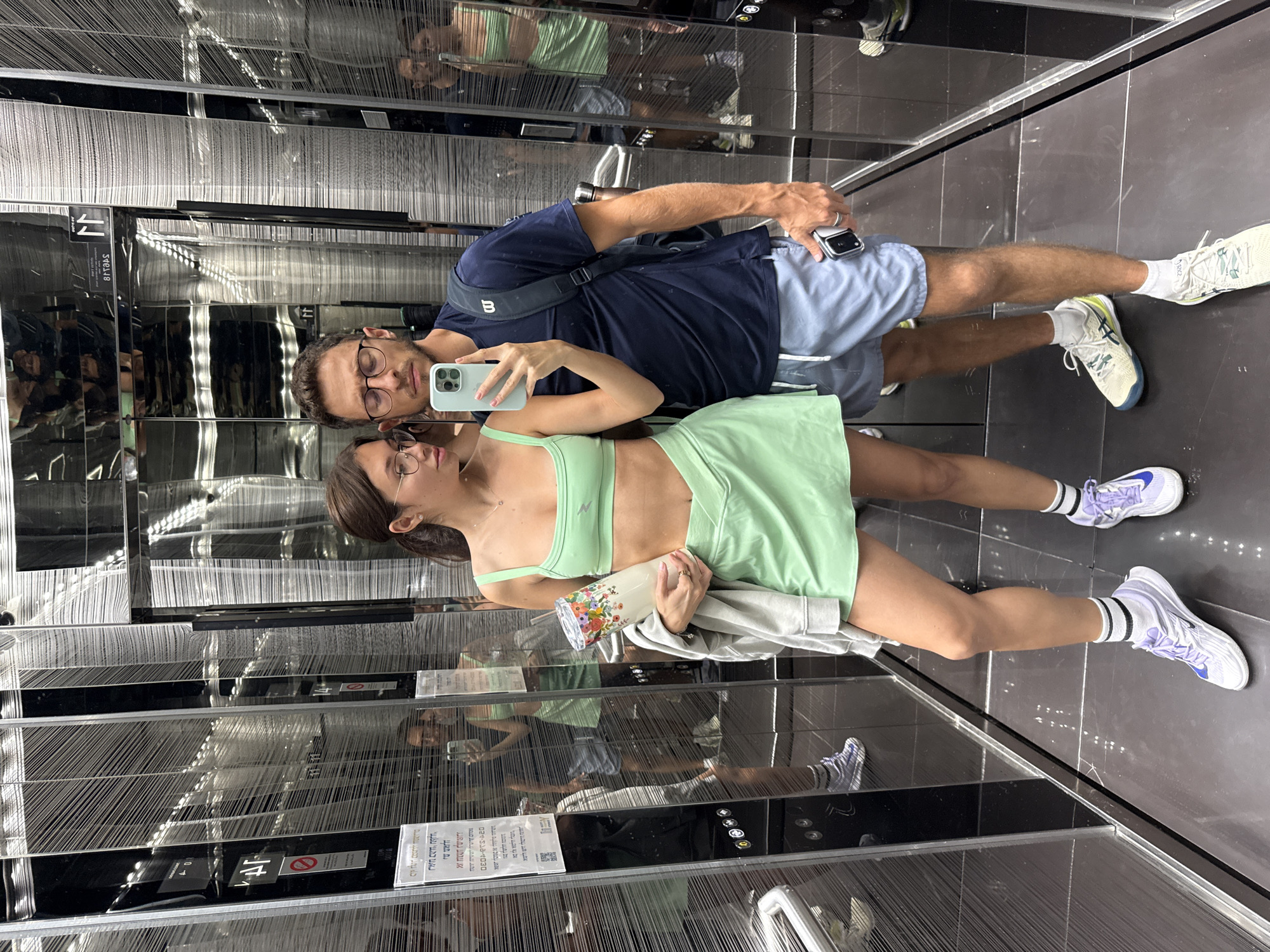 The image shows two people in what appears to be an elevator with mirrored walls, based on the reflections of the subjects and elevator components. The female subject is holding a smartphone to take a selfie, and the male subject is standing beside her, also holding a phone. The woman is wearing a light green athletic outfit and lilac-colored sneakers, and she is also holding a water bottle. The man is wearing a dark blue shirt, blue shorts, and white sneakers.
Signs on the elevator wall contain text in Hebrew, indicating that the elevator is likely located in Israel. The elevator interior consists of mirrored surfaces, black panels, and possibly a textured wall or mesh-like design behind the mirrored surfaces.
While the location can be inferred to be in Israel due to the Hebrew signs, specifying the precise location within Tel Aviv is not possible from the visual information in the image.