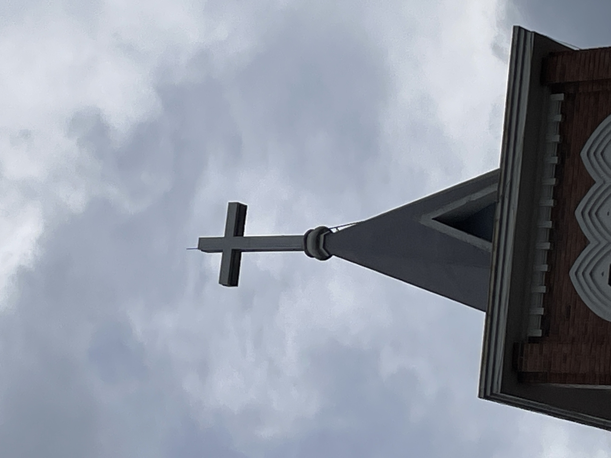 A close-up shot of a Christian cross atop a church steeple against a cloudy sky. The cross is dark gray and appears to be made of metal, with a prominent horizontal bar meeting a vertical one. The steeple is conical and dark gray, with a smaller spire extending upwards from the top of the cross. Below the steeple, the corner of a brick building with white decorative trim is visible. The clouds are dense and gray, suggesting an overcast day. The image lacks any people or discernible activity. The specific location within Lâm Viên - Đà Lạt, Vietnam, is not identifiable from the visual elements alone.
