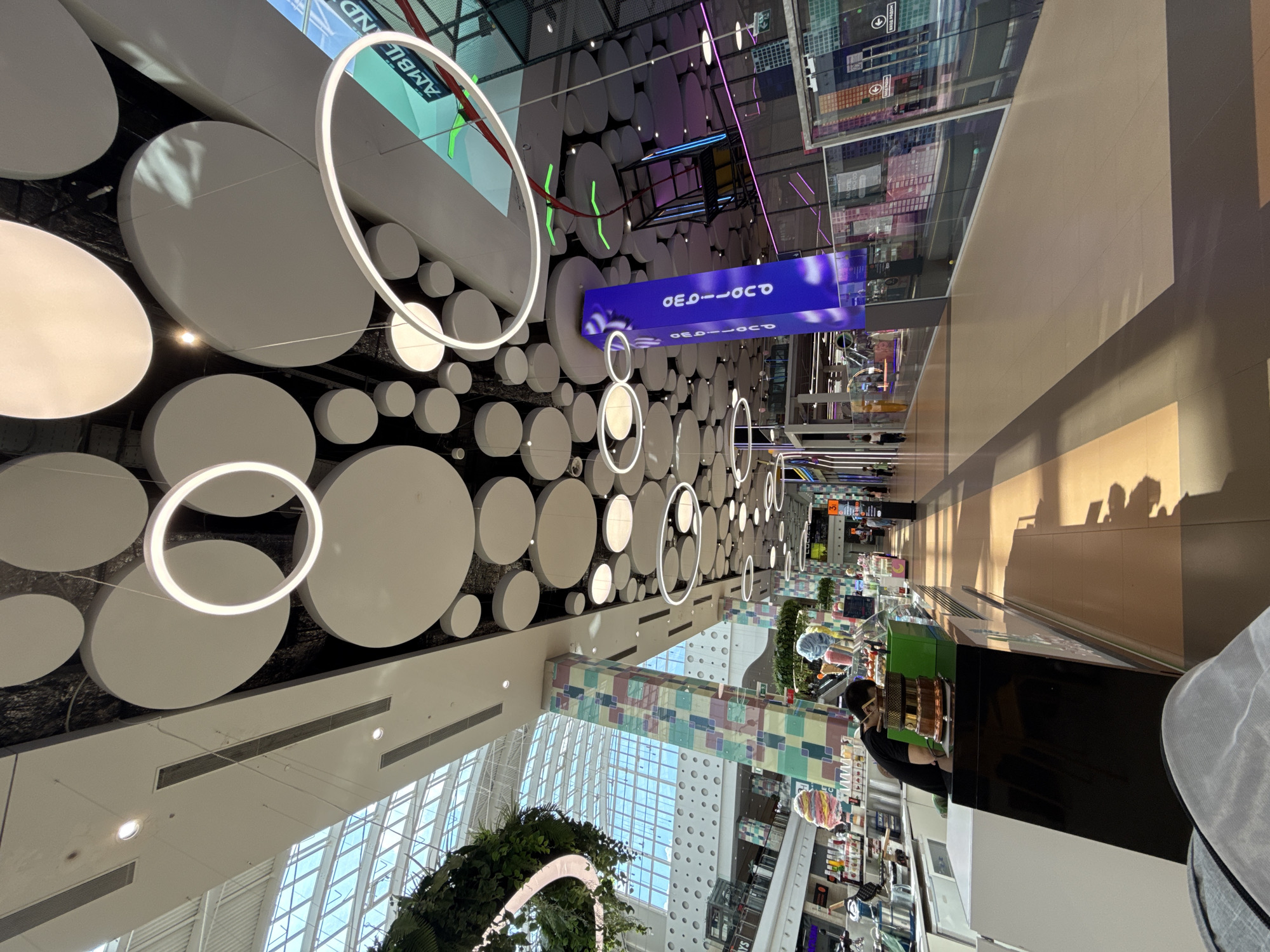 The image shows the interior of a modern shopping mall, likely in Moscow, Russia. The ceiling features a striking design of large, suspended white circles of varying sizes, some illuminated by ring-shaped lights. A colorful, geometric patterned pillar stands prominently on the right side of the image, displaying the word "GEO" in large, white letters against a purple background.
In the foreground, a person wearing a black t-shirt is seen behind a counter, appearing to be an employee at an ice cream or dessert shop. The counter has a black and white facade. Displayed on the counter are various items, including what appear to be desserts and ice cream cones. Further into the mall, there is a walkway with shops on either side, and people are walking through the space. The mall's architecture includes a large glass ceiling that allows natural light to flood the interior, creating distinct shadows on the floor.
In the background, there are escalators and multi-level structures typical of a large shopping center. A sign with the word "AMB" and a green element is visible on the upper right. The overall atmosphere is bright and contemporary, with a focus on modern design elements. The image captures a slice of everyday life within a bustling commercial environment.
