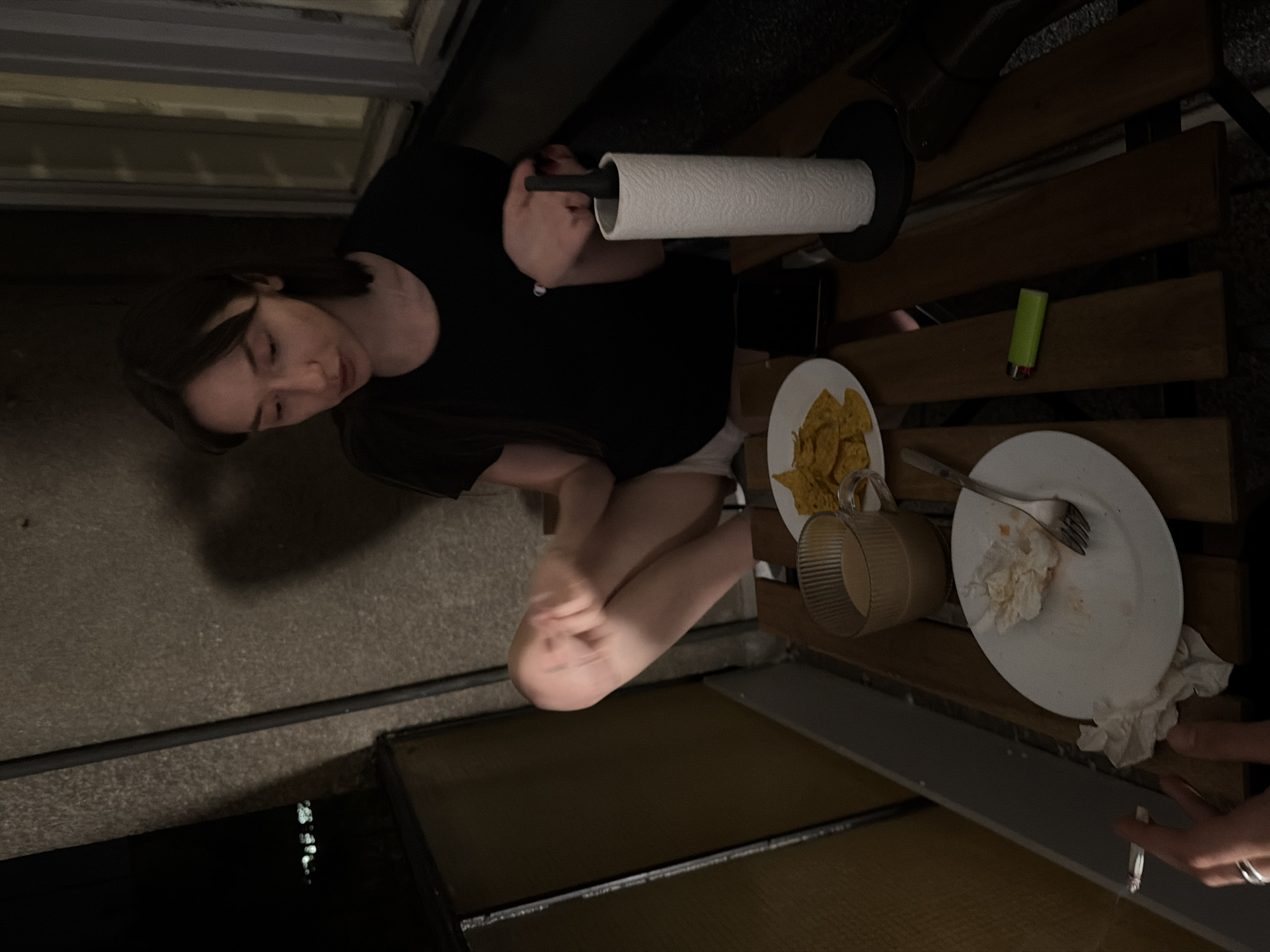 This image captures an outdoor evening scene, likely on a balcony or patio, depicted from an overhead perspective and rotated 90 degrees counter-clockwise. A young woman with dark hair, wearing a black t-shirt and light-colored shorts, is seated on the left side of the frame, looking downwards. Her legs are bent, and her right hand rests near a roll of white paper towels on a stand.
The setting includes a slatted wooden table to the right of the woman. On the table, there is a small white plate with yellow tortilla chips, a clear, ribbed glass mug, and another white plate containing a fork and several crumpled, soiled white napkins or tissues, indicating a recently finished or ongoing meal. A bright green lighter is also visible on the wooden table slats.
In the bottom right corner, part of another person's hand is visible, holding a lit cigarette. The ground beneath appears to be concrete. The dim lighting suggests it is nighttime. No visible text is present in the image.