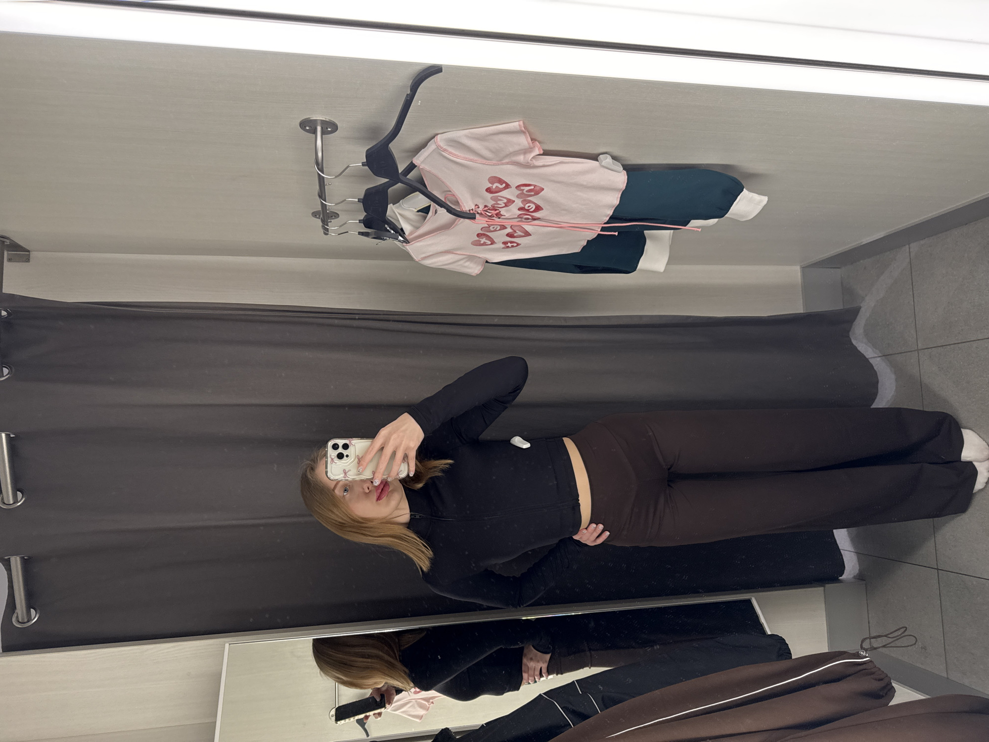 The scene depicts a female individual within a changing room located in the city of Ovoshchnoy, Russia. The subject, a woman with light brown hair, fair skin, red lipstick, and a silver nose ring, is centrally positioned, taking a selfie with a white smartphone held in her right hand. She wears a black, long-sleeved, high-collared cropped top with a zipper, dark brown wide-leg pants, and white socks. Her left hand rests on her hip. A full-length mirror to her left displays her reflection. The changing room features light grey textured walls, a grey tiled floor, and a dark grey curtain with metal grommets pulled to the left. A chrome bar with three black velvet hangers is mounted on the right wall, holding a light pink short-sleeved top with red heart graphics and pink lacing, and a dark green long-sleeved top with white cuffs underneath. A vertical light fixture runs along the far right wall. A brown garment with white piping is partially visible on the floor to the left.