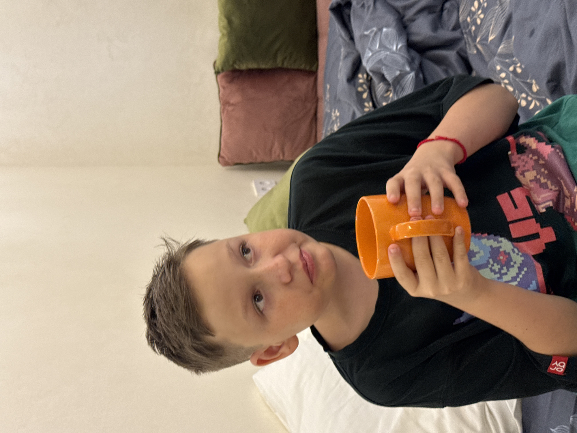 A young boy with short brown hair and freckles is holding an orange mug in his right hand and is looking to his left. He is wearing a dark t-shirt with a colorful graphic design on the front and a green undershirt. He has a red string bracelet on his left wrist. He is sitting on a bed with a white pillow behind him and a dark grey duvet with a floral pattern. In the background, there are two cushions, one dusty rose and the other olive green, against a beige wall. The setting appears to be indoors, possibly a bedroom. There is no visual information in the image that indicates the location is in Sevastopol, Ukraine.