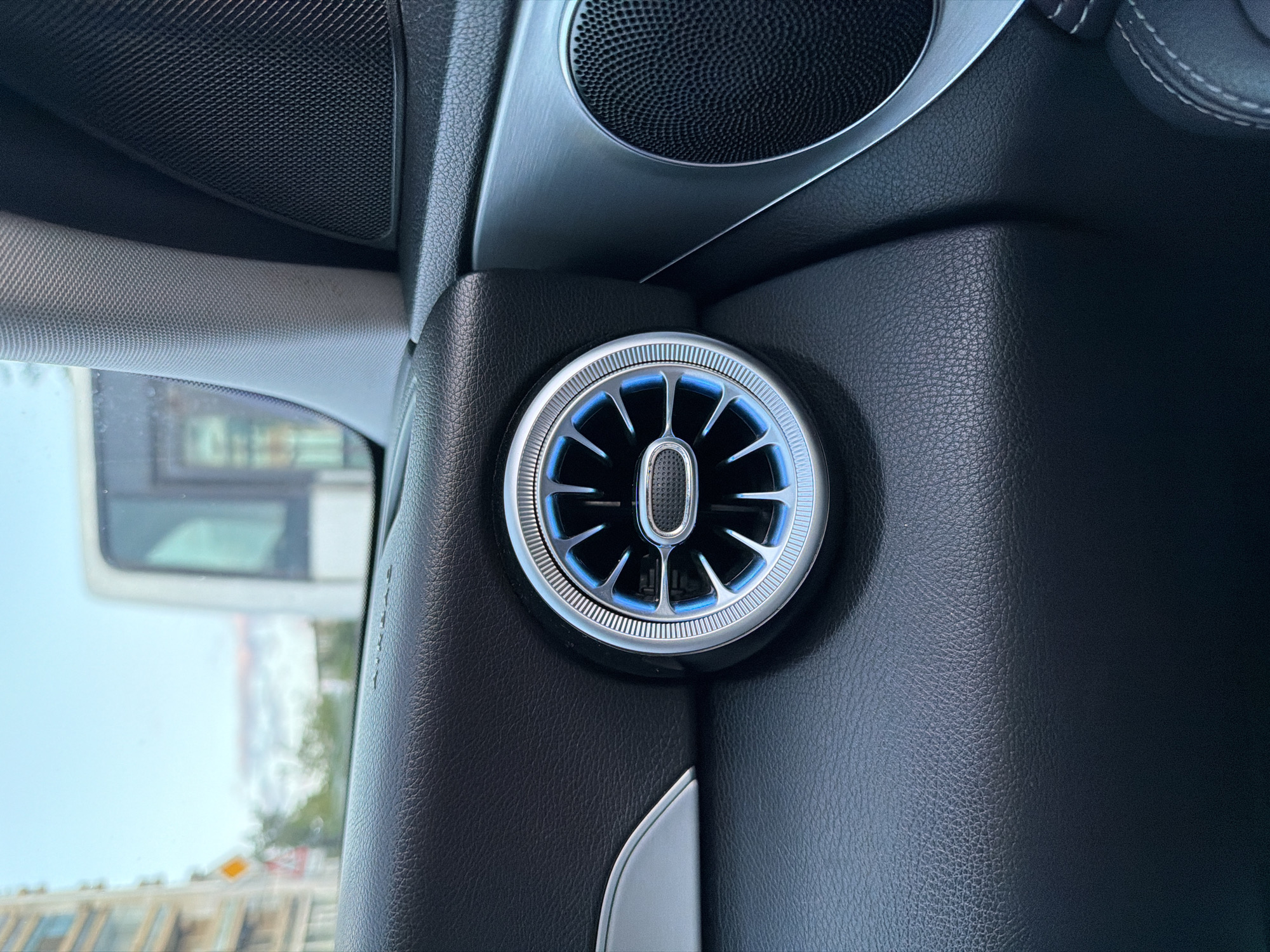 The image captures a detailed view of the interior of a modern car, likely from the passenger side, showcasing the aesthetics of the cabin. The most prominent feature is a circular air vent positioned on the door panel or pillar, distinguished by its metallic silver trim and ambient blue lighting emanating from behind its black fins. The vent's design suggests a contemporary and possibly luxury vehicle.
Above the air vent, a speaker grille made of a finely perforated black material is visible, also encircled by a silver-toned trim piece, blending seamlessly into the car's interior design. The surrounding surfaces are covered in a dark, textured material that appears to be leather or a high-quality synthetic equivalent, exhibiting fine grain patterns and subtle stitching in some areas, indicative of meticulous craftsmanship.
Through the car window, a blurred outdoor scene is visible, suggesting daytime. Glimpses of what appears to be urban architecture, a road, and a large white vehicle (possibly a bus or truck) can be seen. A yellow, diamond-shaped road sign, common in European traffic signage, is faintly discernible in the distance. The light gray fabric of the car's A-pillar or headliner is also present in the upper left portion of the frame. The overall impression is one of a well-appointed and modern vehicle interior, captured in Shumen, Bulgaria. No legible text is visible in the image.