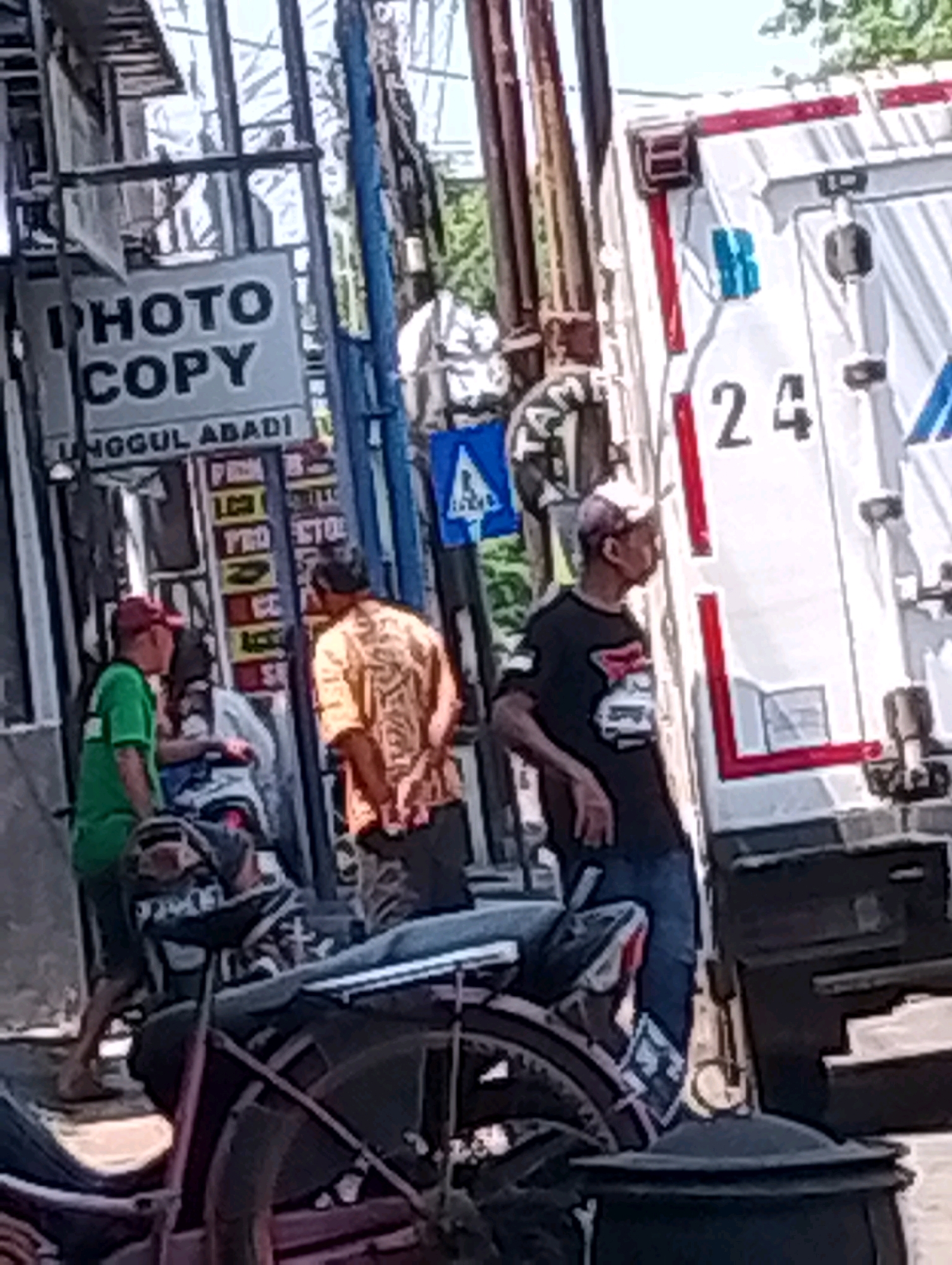 The image captures a street scene in Probolinggo, Indonesia, likely in front of a business. A sign with "PHOTO COPY" is prominently displayed. Several individuals are present. One man in a green shirt and red cap stands near the left side of the frame, facing away from the camera. Another man in an orange patterned shirt stands to the right, looking to the left. A third man, wearing a light-colored cap and a dark t-shirt with a graphic, is on the far right, looking to the left. In the background, a white vehicle, possibly a truck, with the number "24" visible on its side, is parked. A motorcycle is also visible in the foreground, partially obscuring the lower portion of the scene. The environment appears to be an urban setting with a mix of buildings and overhead structures. The overall lighting suggests a bright, daytime scene.