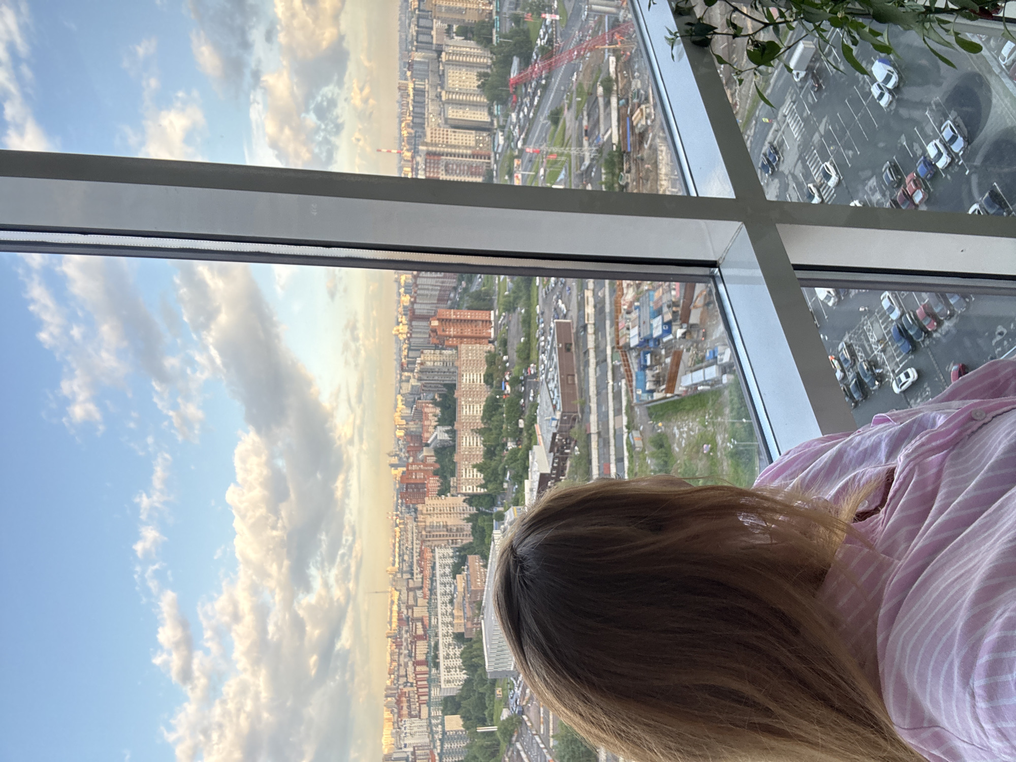 A woman with long, light brown hair, wearing a pink and white striped collared shirt, looks out from behind a large window at a sprawling urban landscape in Pushkin, Russia. The cityscape features numerous apartment buildings of varying heights and architectural styles, predominantly in shades of beige, orange, and grey. Green trees are interspersed throughout the city, and in the foreground, there are signs of ongoing construction with cranes and building materials visible. Below the buildings, a road network is visible with traffic and a parking lot filled with cars. The sky above is a mix of blue with fluffy white and grey clouds, suggesting it is daytime, possibly late afternoon or early evening due to the soft lighting.