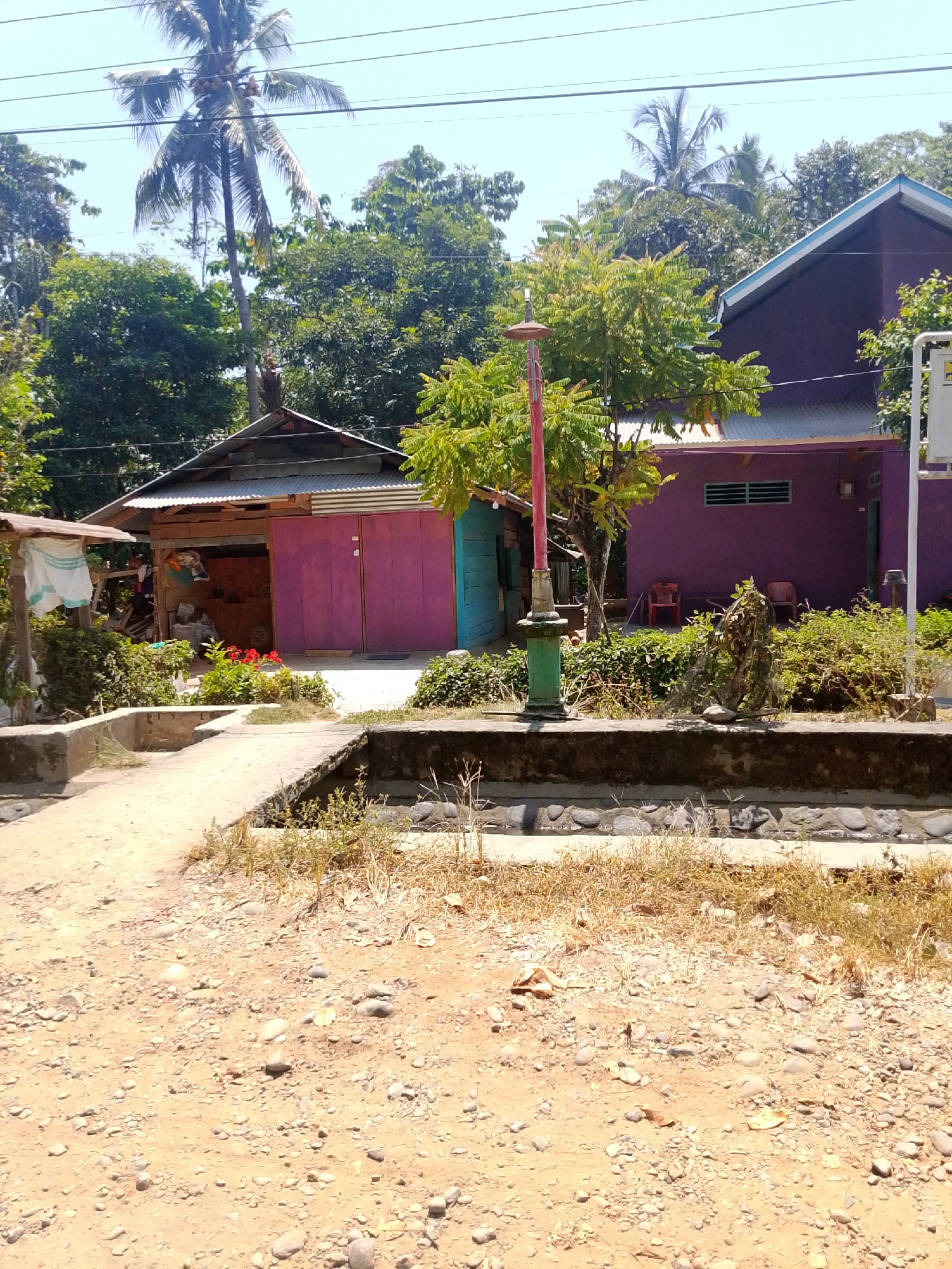 The image depicts an outdoor scene in Se'pon, Indonesia, on a sunny day. The foreground is dominated by a dirt path scattered with small stones and dried vegetation. A low concrete structure runs horizontally across the middle ground, appearing to be a bridge or divider with a small channel below.
Behind the concrete structure, there are two residential buildings. The building on the left is smaller with a corrugated metal roof and wooden walls, featuring vibrant purple double doors and a section of teal-colored wall. To the left of this building, a small, open-sided shelter is visible, with white cloth hanging from a wooden post. Red flowers bloom in front of this structure.
The building on the right is larger and painted a deep purple, with a light blue trim along the roofline. It has horizontal green window blinds. Two red plastic chairs are placed outside this building. A street lamp with a green base and a reddish-pink pole stands between the two buildings, adorned with some foliage.
The background is filled with lush green trees and palm trees, characteristic of a tropical environment. Electrical wires are strung across the top of the image. The overall impression is of a rural or semi-urban residential area in Indonesia. No people are visible in the image.