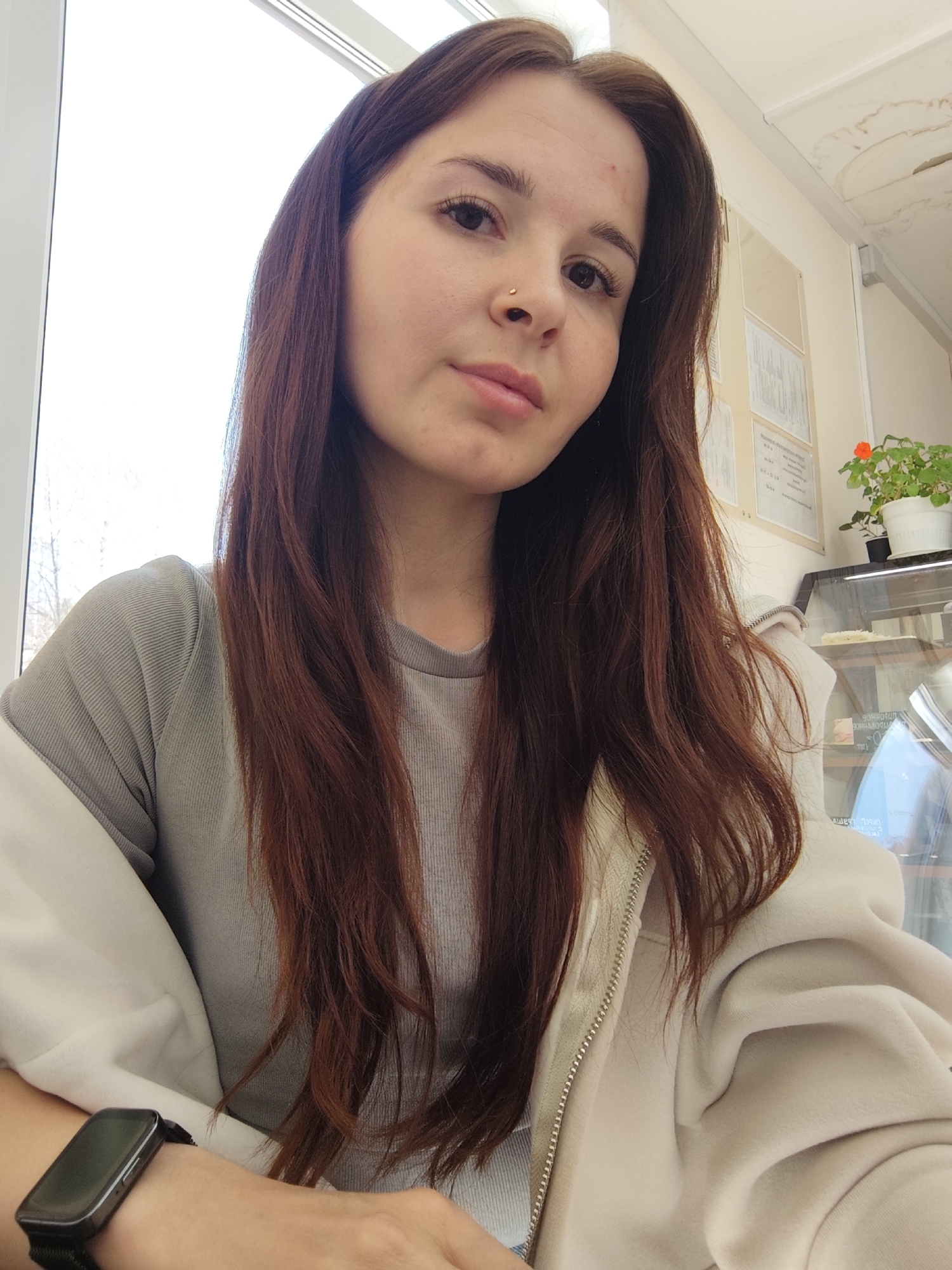 A young woman with long, auburn hair and a nose piercing is posing for a selfie. She is wearing a grey t-shirt and a beige zip-up hoodie. A black smartwatch is visible on her left wrist. The background shows a window on the left side of the frame, a bulletin board, a plant in a white pot with an orange flower, and a display case with food items. The picture may have been taken in a café or similar establishment in the city of Chaikovsky, Russia. The ceiling shows signs of water damage.