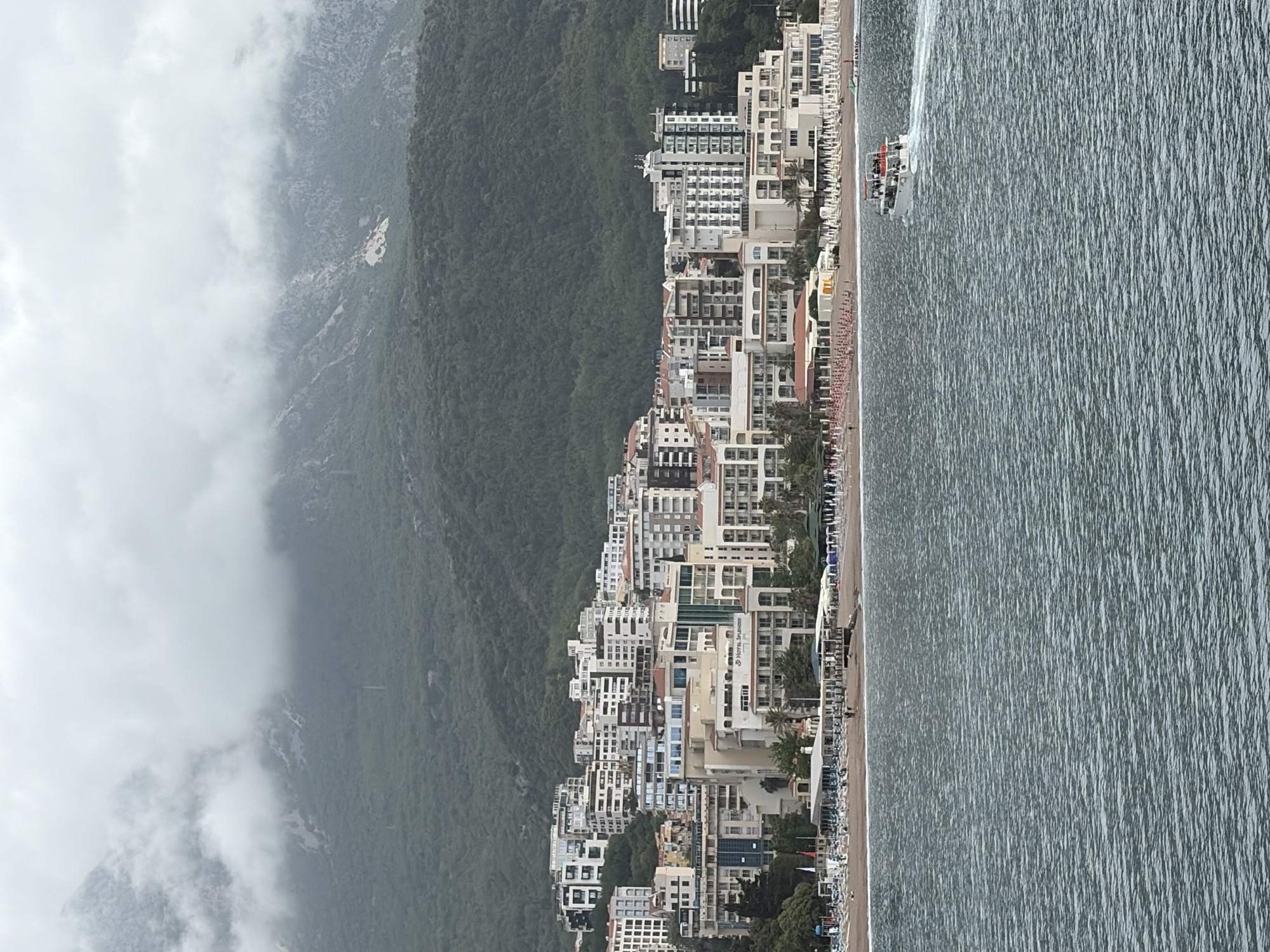 The image captures a coastal view of Budva, Montenegro. The scene is dominated by a line of multi-story buildings and hotels clustered along the shoreline, fronting a beach with rows of beach chairs and umbrellas. Adjacent to the buildings is a densely vegetated hillside that rises up into a fog-covered mountain range. To the right of the buildings and beach, the waters of the Adriatic Sea extend into the horizon. A boat is visible in the water near the shore, leaving a wake behind it. The overall atmosphere is overcast, with subdued lighting.