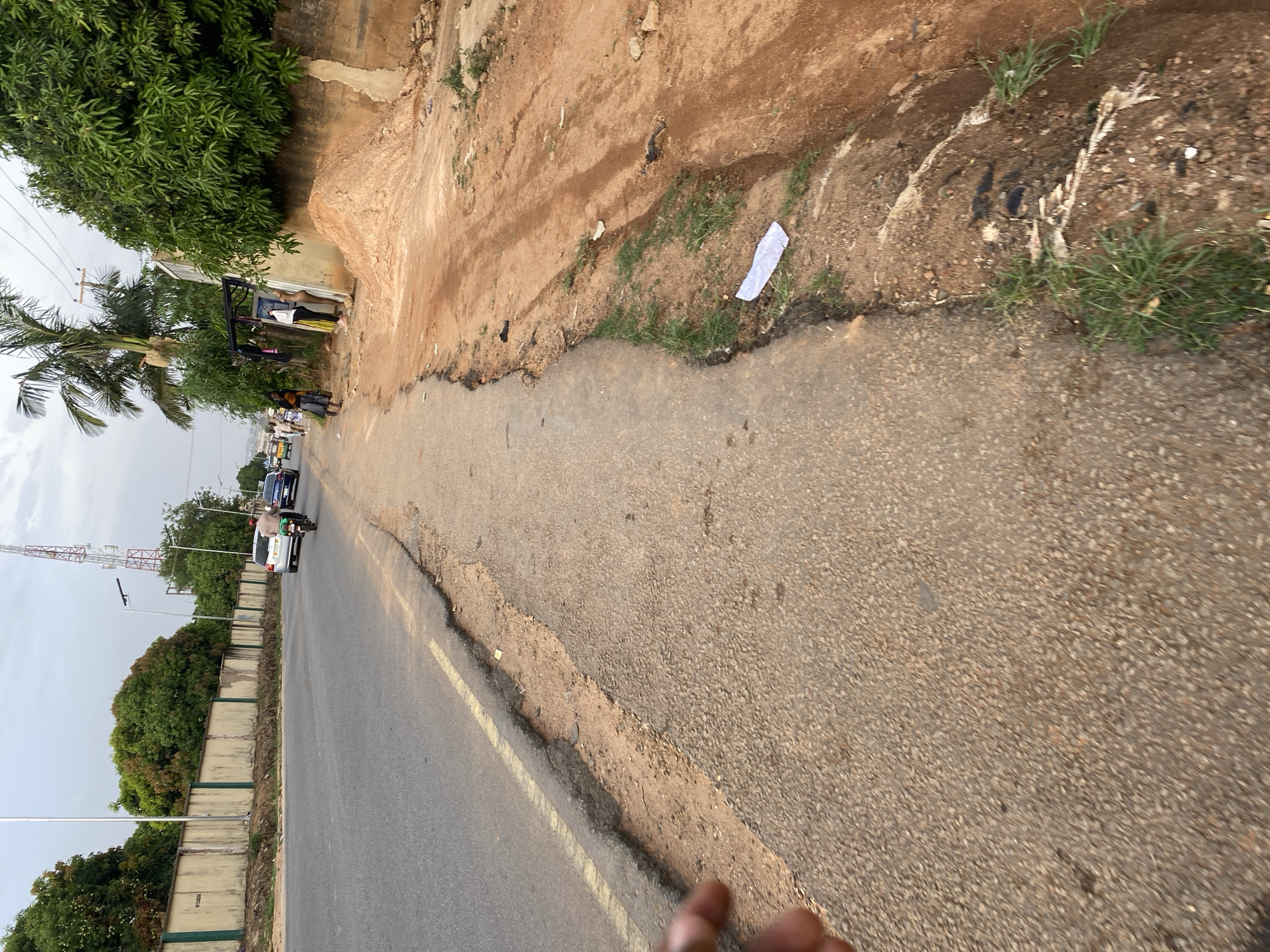 The image depicts a street scene in Bukuru, Nigeria. The foreground shows a rough, paved road with a yellow painted line and a dirt shoulder. Beyond the shoulder is a raised dirt embankment with sparse vegetation. In the background, there are several people gathered near some buildings. A palm tree and other trees are also visible. There are vehicles on the road, including cars and a motorcycle. A tall telecommunications tower is seen in the background. The sky is overcast.