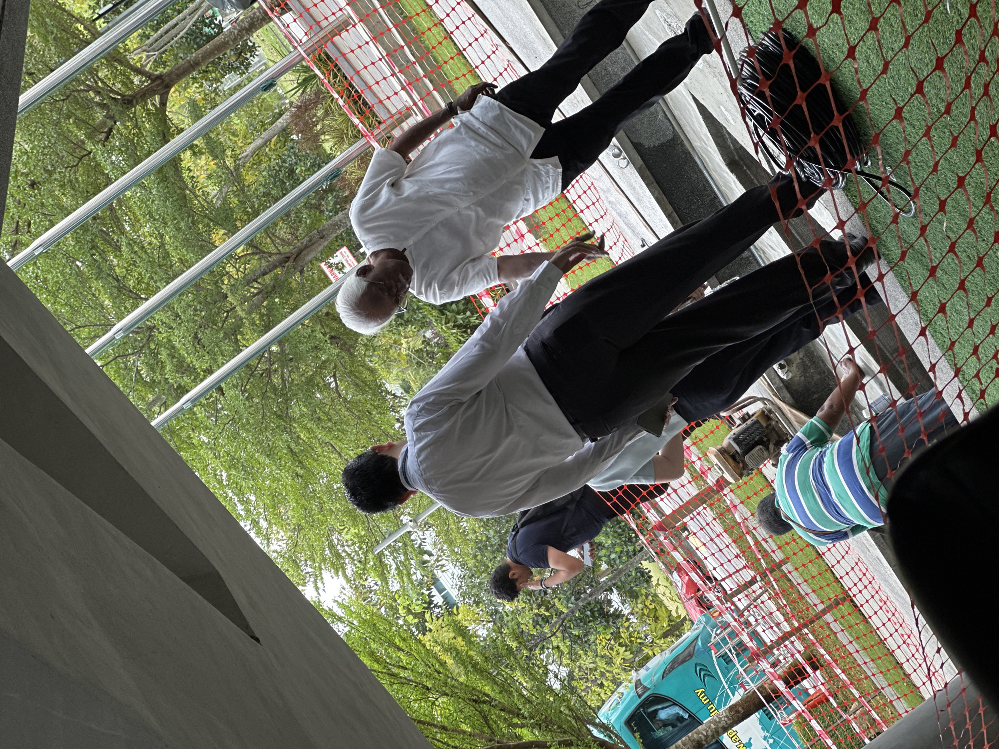 The image depicts an outdoor scene in Kuala Lumpur, Malaysia. Two men in light-colored shirts and dark pants stand on a paved surface next to a red safety net. A third man wearing a blue and green striped shirt is partially visible near the edge of the frame. A fourth man bends to inspect something near the curb. A light blue vehicle with visible writing on its side is parked on the street behind a tree, which hints at the urban location of Kuala Lumpur. Overhead are tree branches and steel beams or supports.