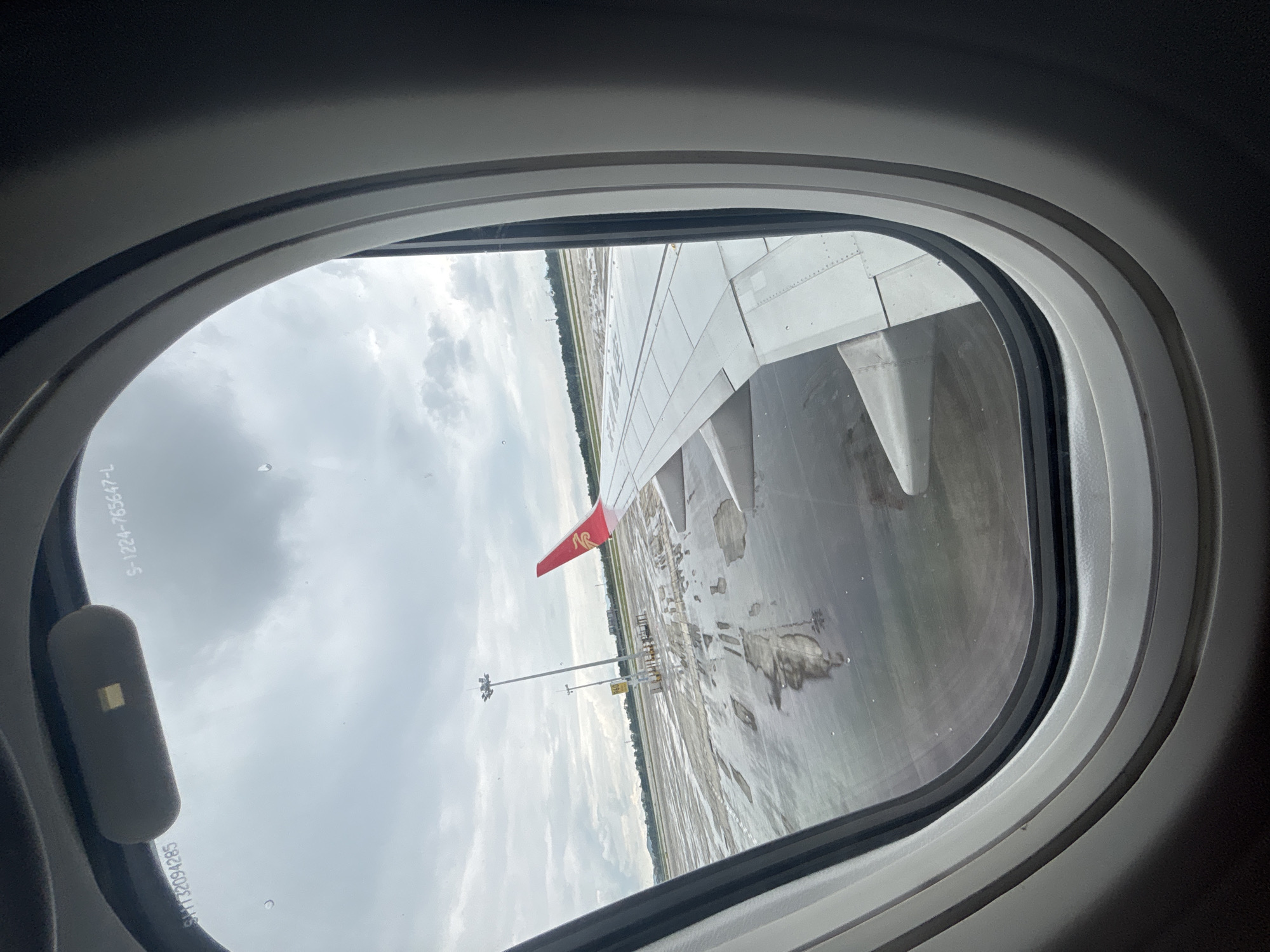 The image shows a view from an airplane window, looking out at an airport tarmac under a cloudy sky. The tarmac appears wet, with puddles reflecting the overcast conditions. A portion of the airplane's wing, with a red logo on its tail, is visible on the right side of the frame. In the distance, there are tall light poles and some buildings. The window frame of the aircraft is visible around the edges of the image. There are some markings on the window itself, including "S-1224-765647-L" and "SH132094285". The scene suggests an airport environment, possibly during or after rain. Given the context of Baiyun District, China, this is likely the Baiyun International Airport (CAN).