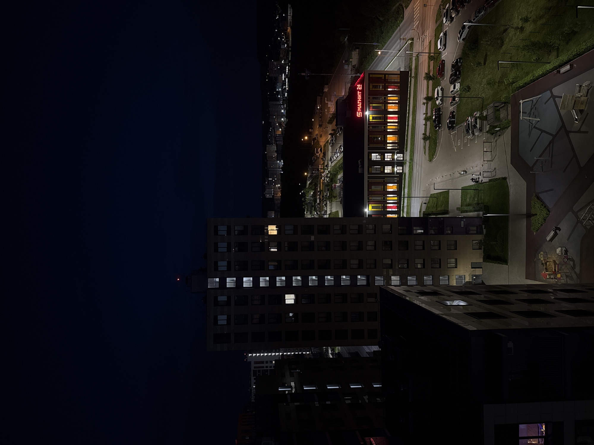 The image depicts an aerial view of a residential and commercial area at night, likely in the city of Perm, Russia. Dominating the foreground are two tall apartment buildings, one a darker, more angular structure on the left, and a lighter-colored, rectangular building on the right with numerous illuminated windows. In the midground, a low-rise building with a red, illuminated sign reading "МАГНИТ 24" is visible. This building appears to be a retail establishment, possibly a supermarket, with colorful illuminated windows and a parking lot in front of it filled with several cars. A paved road with streetlights runs alongside the retail building and a parking lot. Further into the background, a sprawling cityscape with more buildings and lights stretches towards the horizon under a dark night sky. The overall scene suggests a modern urban development.