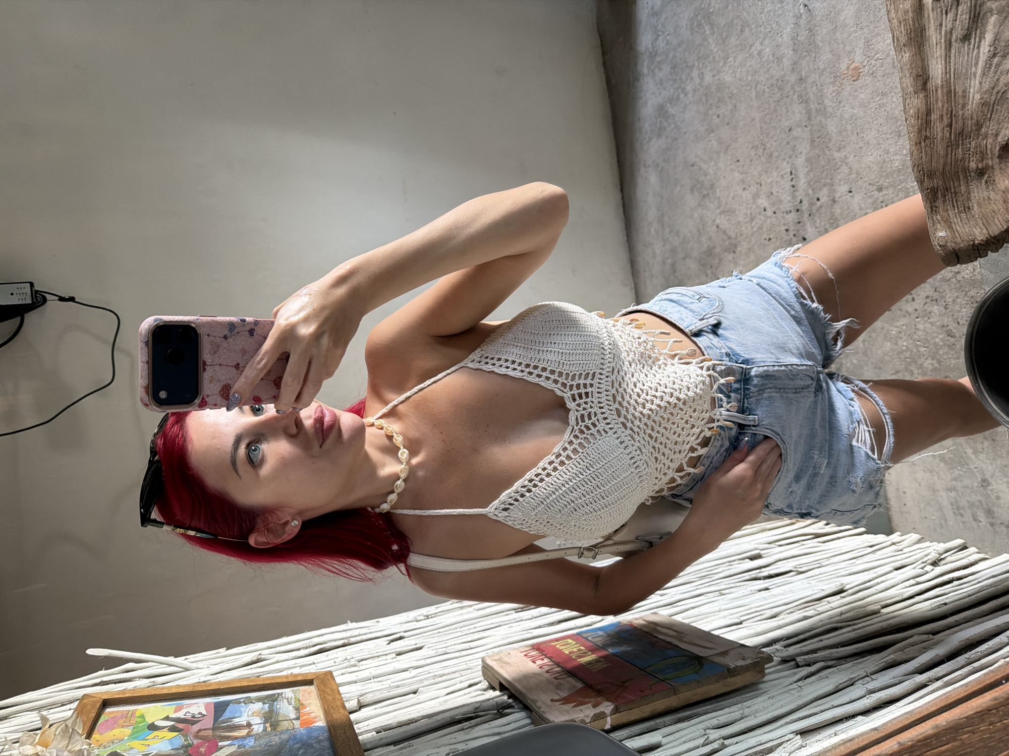 A young woman with vibrant red hair is taking a selfie in an indoor setting. She is wearing a white crocheted halter top with a pearl necklace and ripped denim shorts. Her left hand is holding a pink-cased smartphone up to take the photo, while her right hand is tucked into the pocket of her shorts. She is looking at the phone's screen with a neutral expression.
The background appears to be a room with a light-colored wall and a concrete floor. To the right of the woman, a piece of weathered wood is visible, and to the left, a framed picture with abstract art is resting on a surface made of white, bundled reeds or branches. The lighting suggests it is daytime, with natural light casting shadows. The overall impression is casual and relaxed.
There is no visible text in the image. The context provided indicates the location is Kuta, Indonesia.