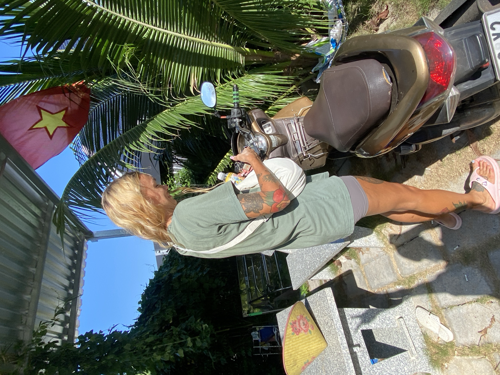 A blonde woman with tattoos on her arms and legs stands next to a brown and gold scooter in Mũi Né, Vietnam. She is wearing a green t-shirt and gray shorts. The woman holds a white bag and a bottle of water. A Vietnamese flag is visible on a pole behind her, partially obscured by palm fronds. The scene is outdoors, with a tiled ground, a stone bench, and lush greenery. A yellow conical hat with floral designs rests on the bench. The scooter's taillight is red and its handlebars have green grips.