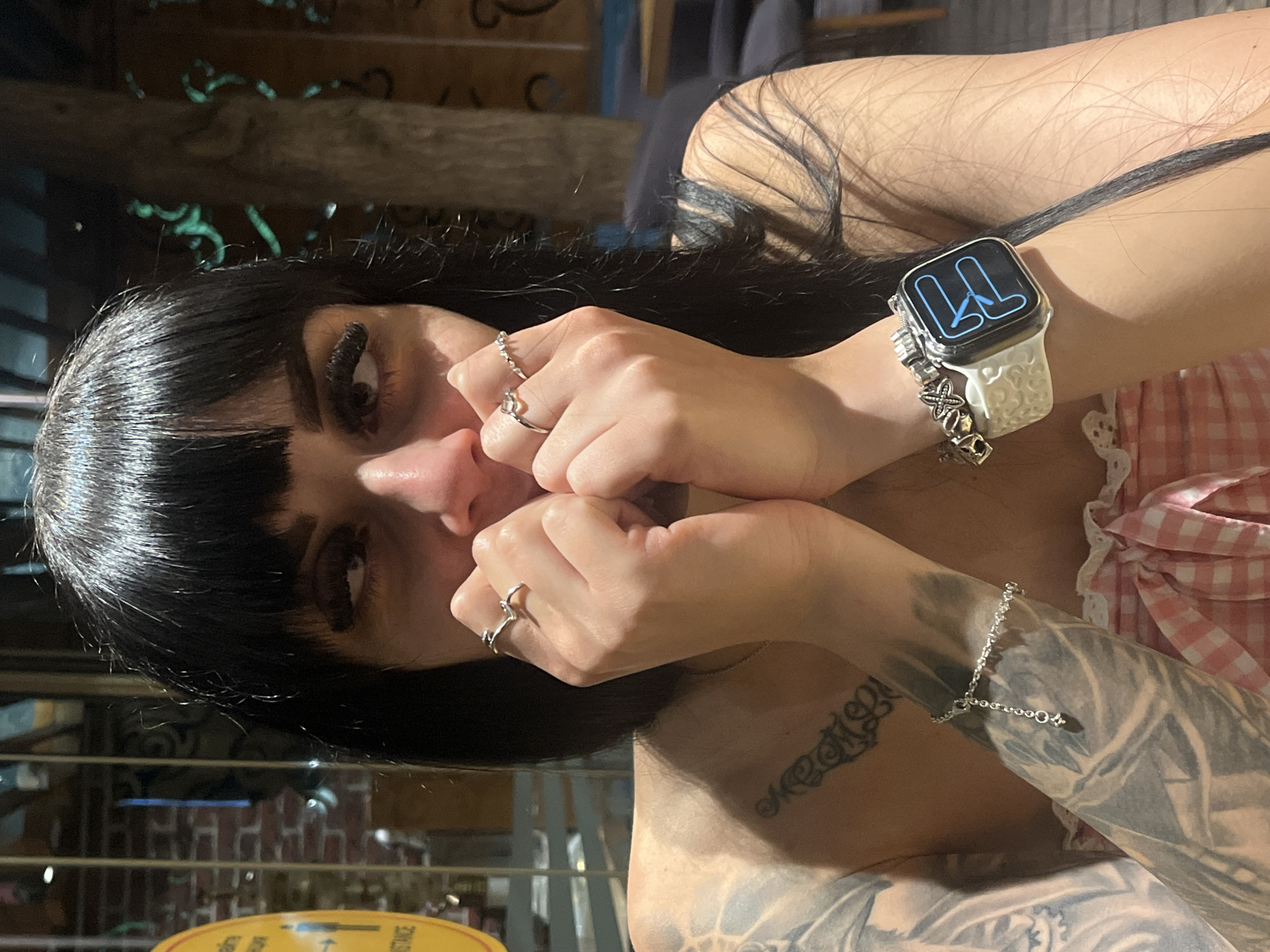A woman with dark hair and bangs is sitting at a table indoors, possibly a restaurant or cafe, with a brick wall and decorative tree visible in the background. She is wearing a pink and white checkered top. Her hands are raised to her face, covering her mouth and nose, with her fingers intertwined. She is wearing several rings on her fingers and a bracelet on her left wrist, along with a smartwatch. She has extensive tattoos on her left arm and chest. The image is a close-up, focusing on her face and upper body. No specific location in Plovdiv, Bulgaria, can be determined from the image.