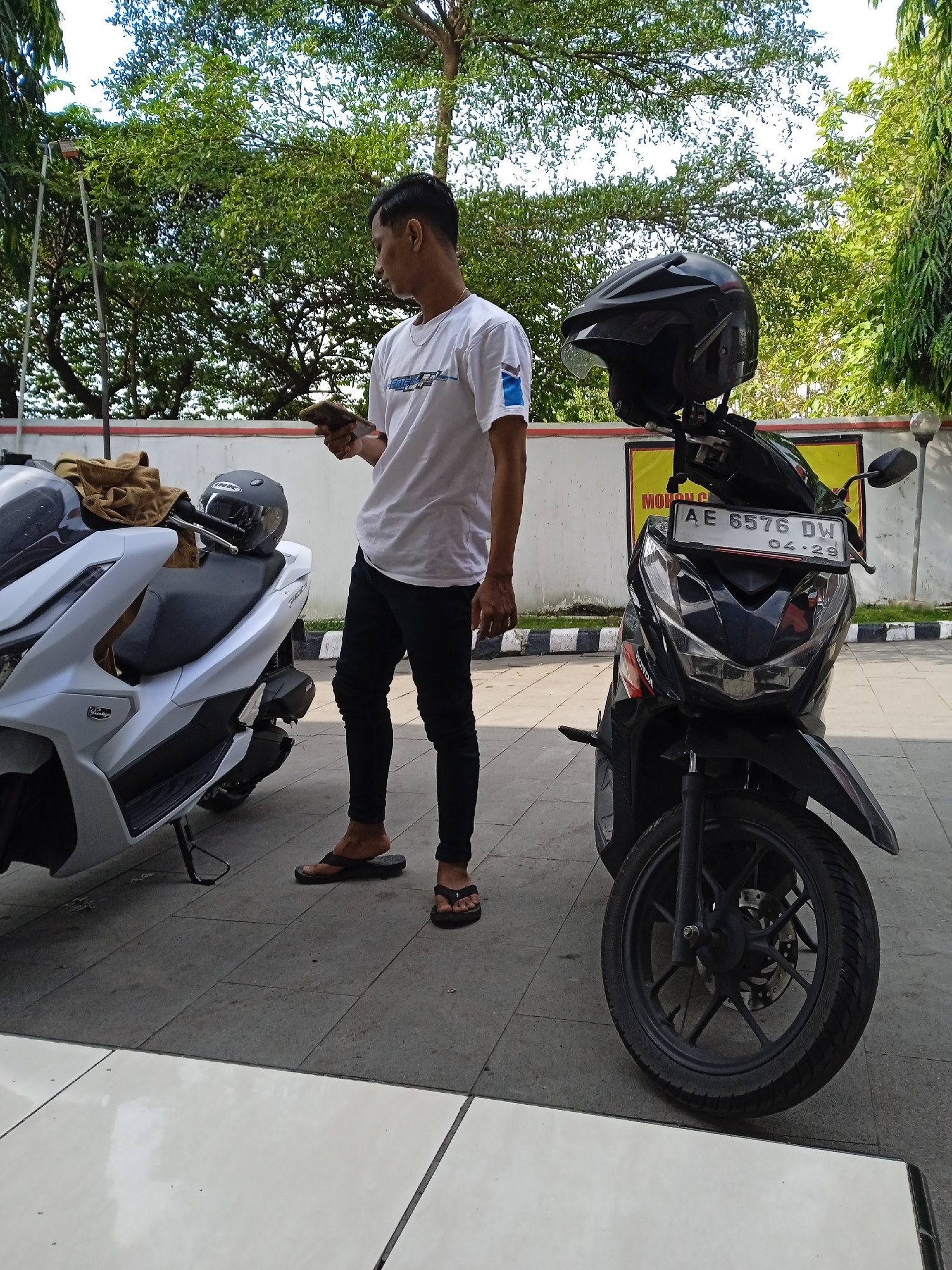 A man stands between two motorcycles in Katonsari, Indonesia. He is facing left, holding a mobile phone. The motorcycle on the left is white with a helmet on the seat and a brown bag attached. The motorcycle on the right is black, with a black helmet strapped to it and a license plate reading "AE 6576 DW 04-29". The man is wearing a white t-shirt, black pants, and flip-flops. The ground is paved. Behind the motorcycles is a low wall and trees.